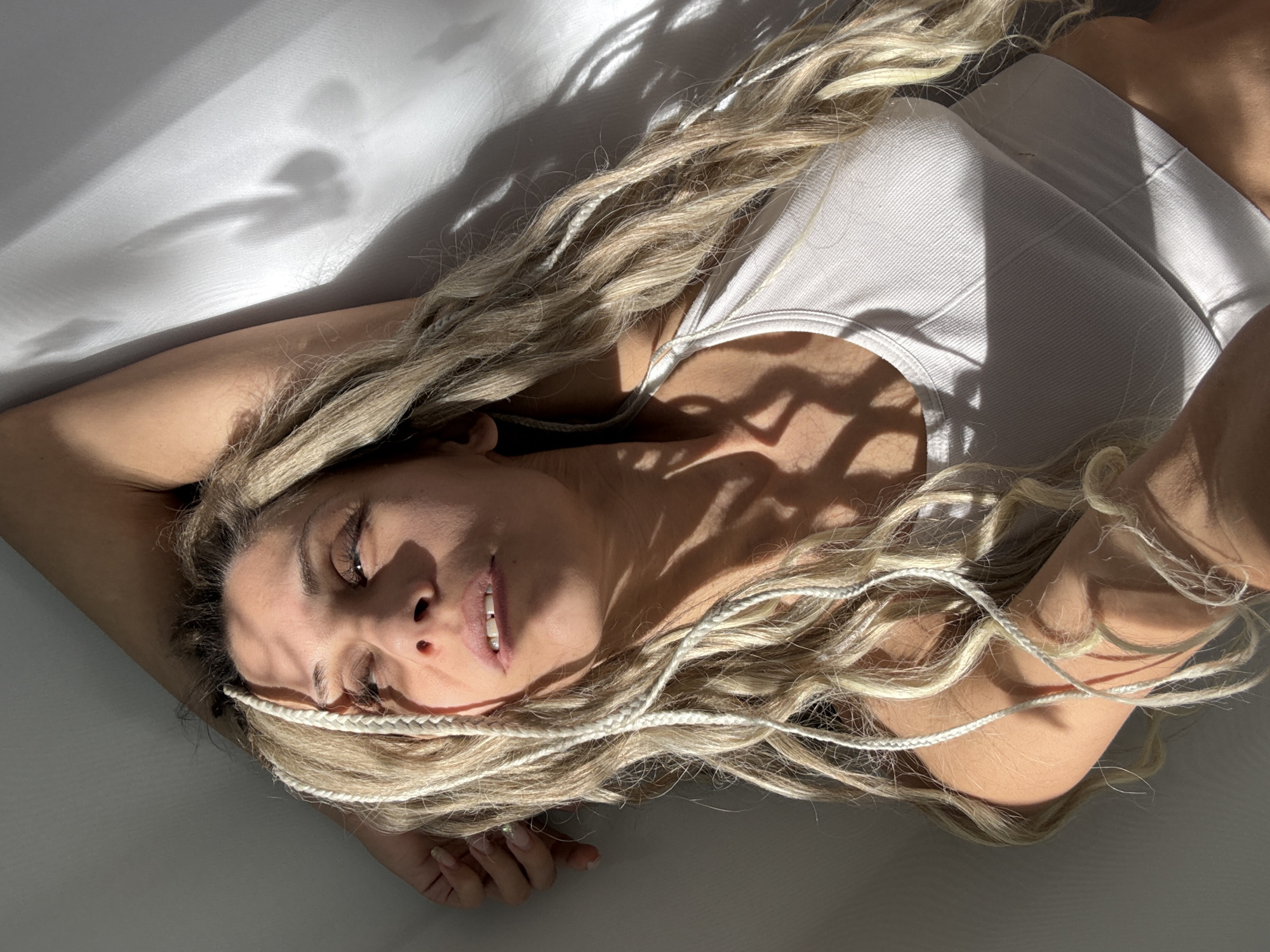 An overhead shot captures a fair-skinned woman with long, voluminous blonde hair, adorned with white braided extensions, lying on a light-colored surface, possibly a bed or floor, in what appears to be an indoor setting. She is wearing a light-colored, ribbed tank top. Her head is slightly tilted, and her arms are relaxed, with one arm extending above her head. She looks towards the camera with a soft, slight smile and a serene expression, her eyes partially open.
The scene is dramatically lit by direct sunlight, casting intricate and prominent shadows across her body and the surrounding surface. These shadows, possibly from a window frame or foliage outside, create a dappled effect, adding depth and texture to the image. The strong contrast between highlights and deep shadows suggests it is daytime, likely morning or late afternoon when the sun is at an angle. The overall mood is calm and introspective. No visible text is present. The location is Rusanovo, Russia.