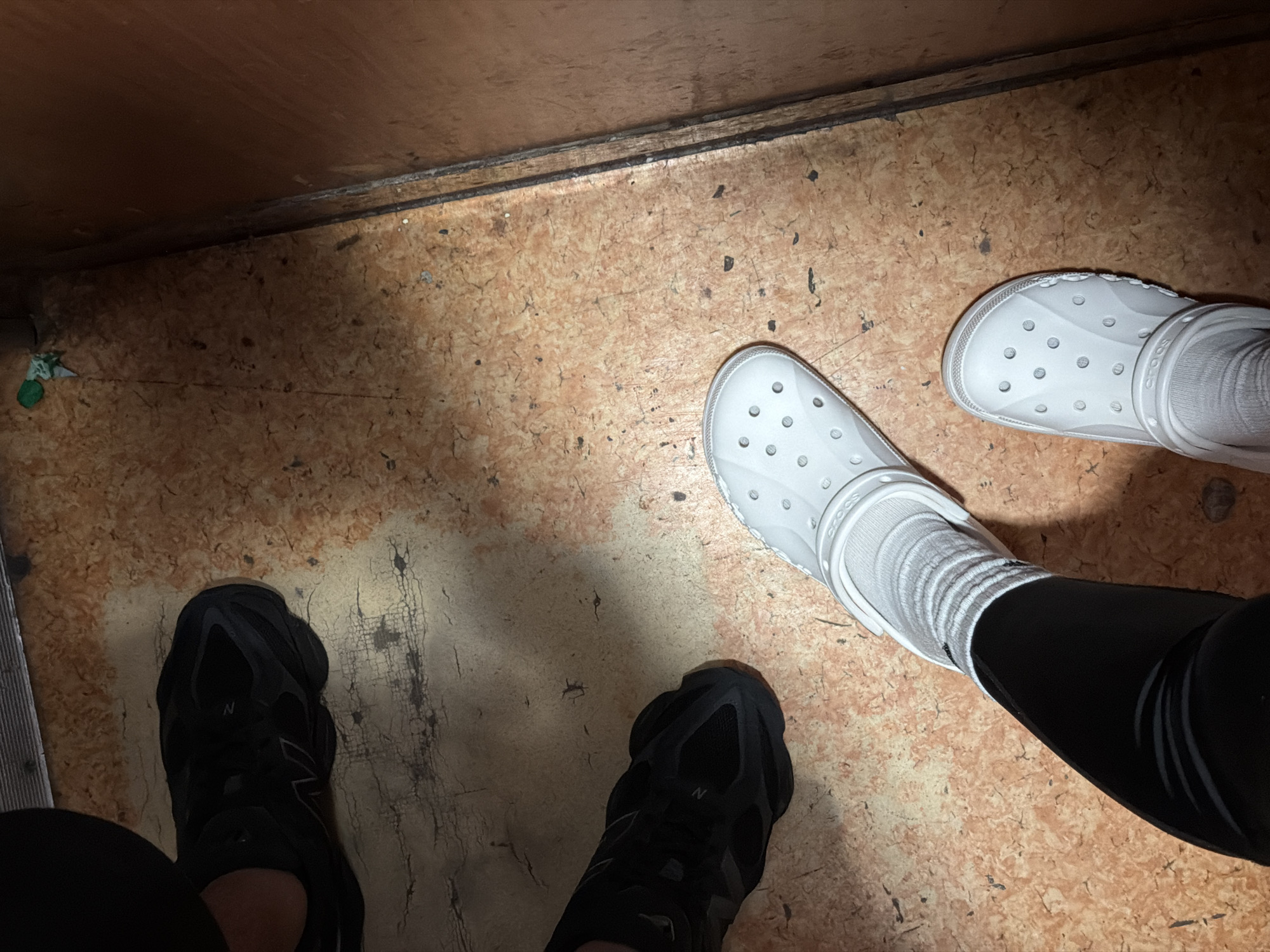 The photograph depicts two pairs of feet on a mottled, light brown floor. The pair on the right wears white Crocs with white socks and black leggings. The pair on the left wears black New Balance sneakers with black leggings. The floor is adjacent to a wall or ledge, indicating an enclosed space. The location is not evident in the image. Therefore, the city of Lovech, Bulgaria, cannot be confirmed as the setting.