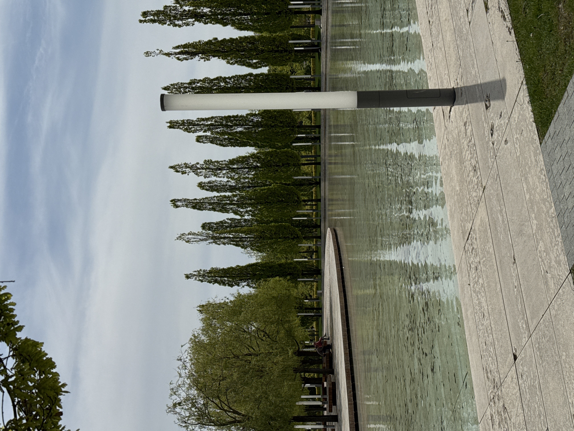 An outdoor landscape scene depicts a serene park or public garden during the daytime under an overcast or partly cloudy sky. A long, narrow body of water, resembling a reflecting pool or ornamental canal, dominates the left and central areas of the frame. Along the far edge of this water, a uniform line of tall, slender trees with dense green foliage creates a structured backdrop, their reflections subtly visible on the water's surface, which also shows gentle ripples.
In the foreground, a modern lamp post with a white, cylindrical light fixture and a dark gray base stands prominently to the right of the water, casting a distinct shadow on the adjacent light-colored, paved walkway. This paved surface runs along the right side of the water, extending into the distance. Beyond the walkway and in the further background, patches of green grass are visible. A large, leafy tree with lush green foliage occupies the bottom-left corner, its branches extending into the frame. In the mid-ground, near the water, a faint figure or object with what appears to be a red bag is visible on a circular wooden platform or seating area.
No specific activity or event is clearly taking place; the scene conveys a sense of calm and order. There is no visible text.