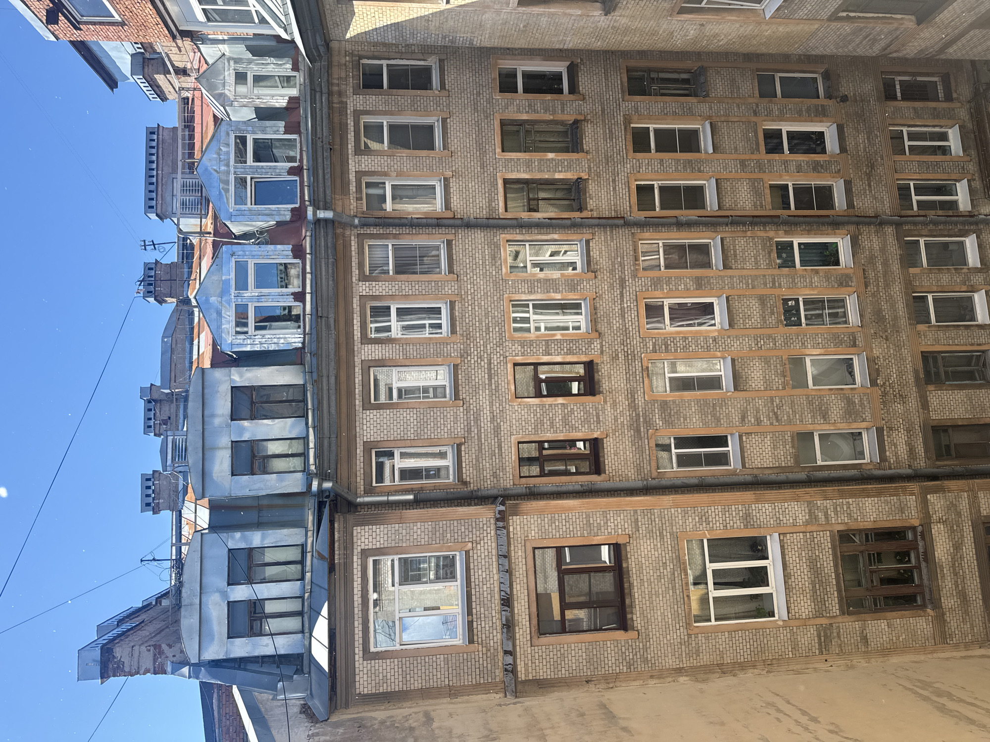 This is an exterior, eye-level shot of a historic courtyard building in Saint Petersburg, Russia, bathed in bright sunlight. The building is constructed of light-colored brick with decorative terracotta trim around the windows and vertically along the facade. Multiple floors of windows are visible, many with dark wooden frames. Some windows have white frames and blinds or curtains. The roofline is complex, featuring dormer windows with metal cladding, chimneys, and what appear to be some modern rooftop additions. Electrical wires crisscross the sky above the building. The overall impression is of a classic Saint Petersburg residential building, likely an apartment block, situated in a tight urban courtyard. No people are visible in the image.