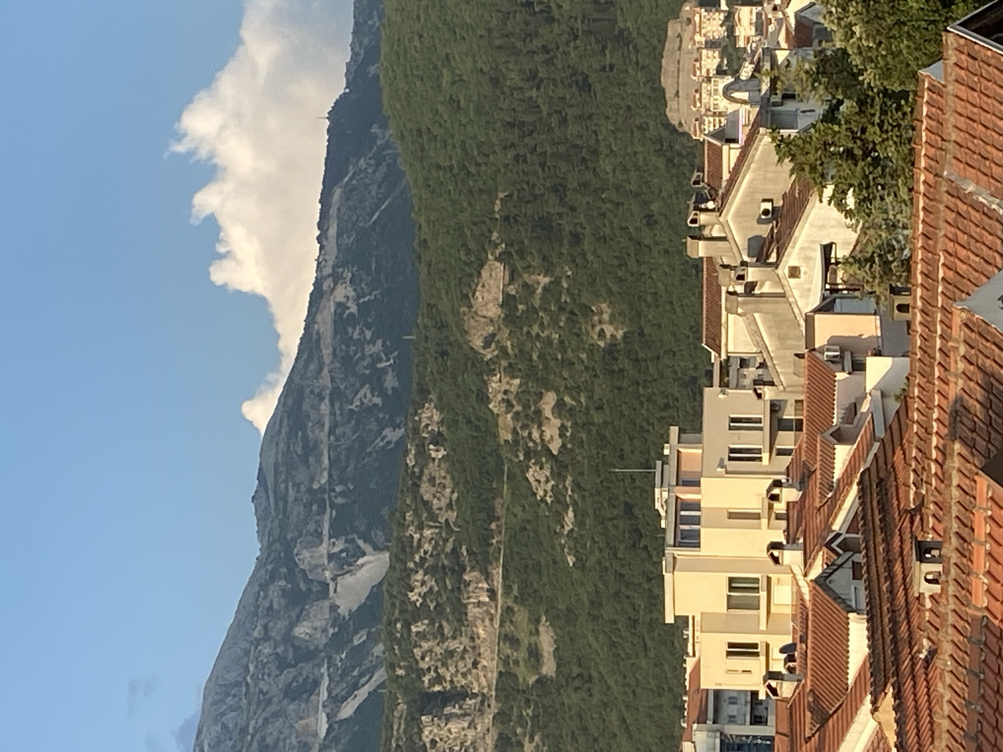 The image shows a cityscape in Budva, Montenegro, with the prominent grey rock and vegetation-covered mountain in the background on the left. Several residential buildings with red tiled roofs and tan walls are visible on the right side of the image. Patches of green trees are visible on the roofs. A clear sky is observed at the top left corner, with some clouds next to the mountain's peak.