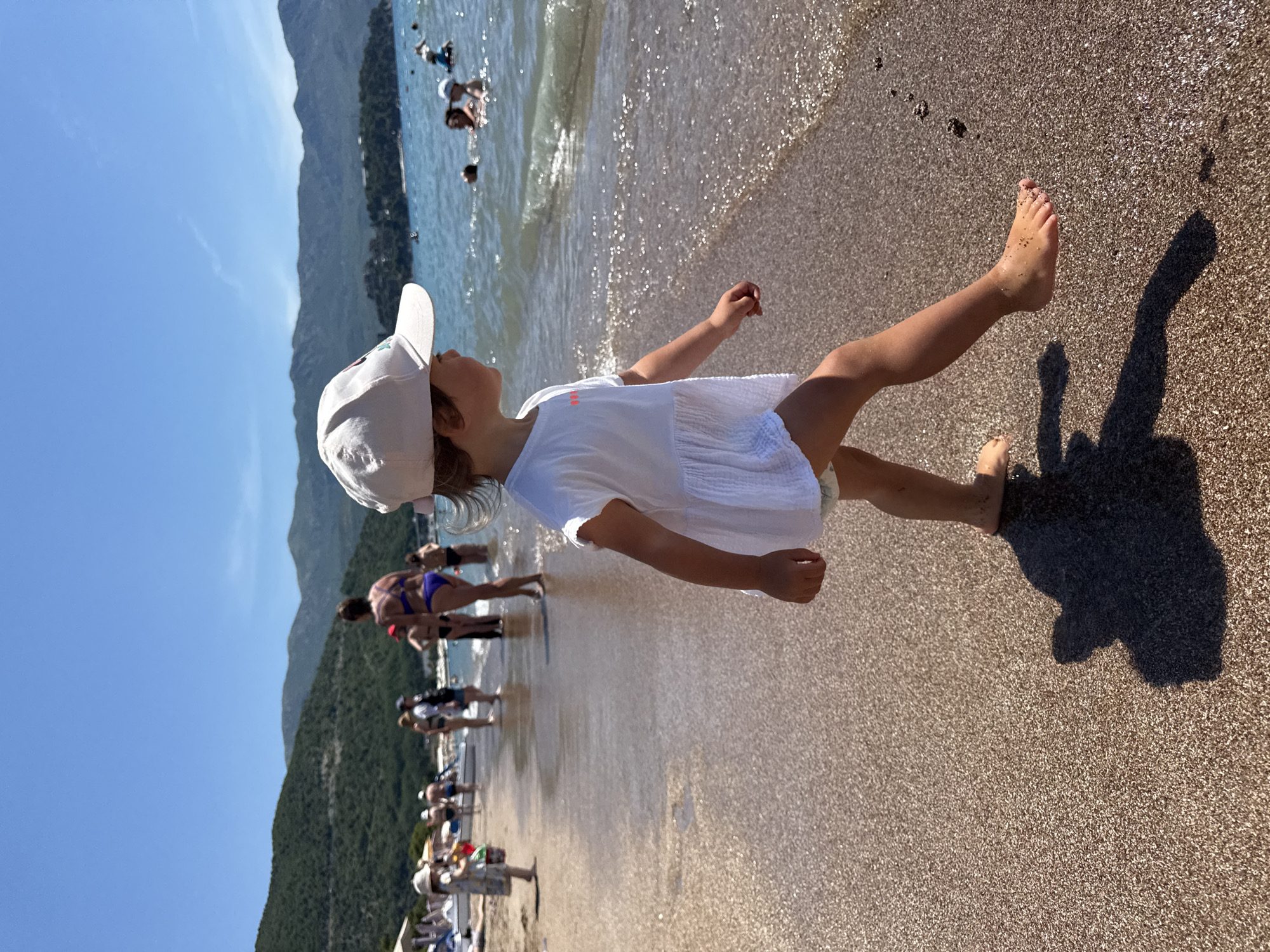 In the city of Budva, Montenegro, on a sunny day at the beach, a young child wearing a white cap and dress walks along the wet sand near the water's edge. The child's shadow is cast on the beach. In the background, several people are in the water, and a few others are standing on the shoreline. Mountains and a clear blue sky form the backdrop.