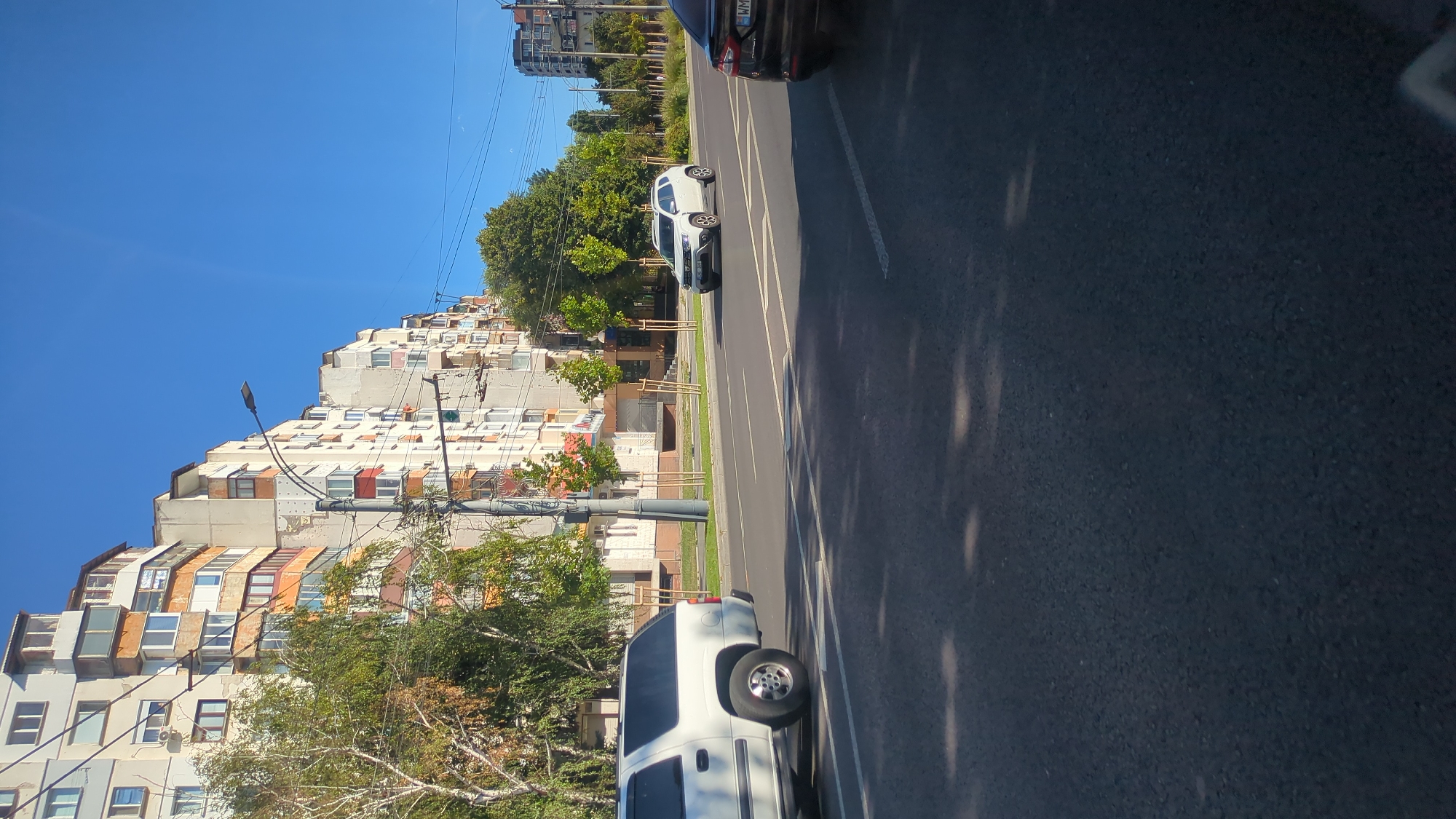 The image depicts a street scene in Chișinău, Moldova, on a clear, sunny day. The focus is on a multi-lane road with several vehicles and a row of apartment buildings lining the left side.
In the foreground, the asphalt road surface is visible, with white lane markings. The sunlight casts dappled shadows on the road.
On the left side of the road, a white SUV is parked or moving slowly. Further down the street, a white Dacia Duster is driving in the right lane. A dark-colored car is partially visible in the right lane, closer to the foreground.
The apartment buildings are tall, with multiple stories and balconies. They appear to be older Soviet-era construction, with some showing signs of wear and tear. The architecture includes numerous windows and varied balcony designs, some with awnings.
Trees with green foliage line the sidewalk in front of the buildings, providing shade. Streetlights are also visible along the road. In the background, more buildings and trees are visible, suggesting a residential or mixed-use urban area. The sky is a vibrant, clear blue.