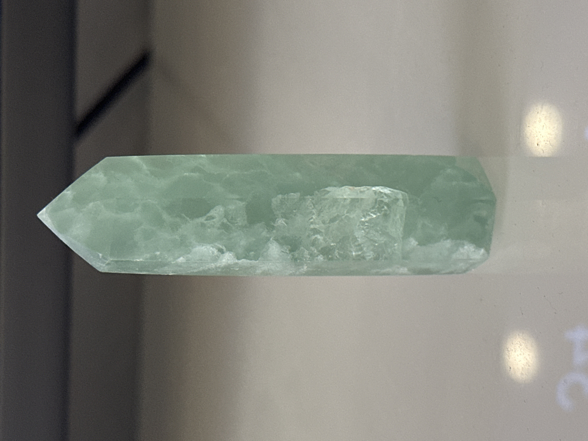 A green, pointed crystal tower rests on a light-colored surface. The background is dark and out of focus, with a hint of horizontal lines suggesting a shelf or furniture. The crystal exhibits internal fractures and variations in color, ranging from pale green to a more translucent shade. There are reflections on the surface beneath the crystal, one appearing as a bright circular spot and another as faint, blurry white shapes. The image is a close-up shot, focusing entirely on the crystal and its immediate surroundings. There is no indication of the city of Nevinnomyssk, Russia, or any other geographical location.