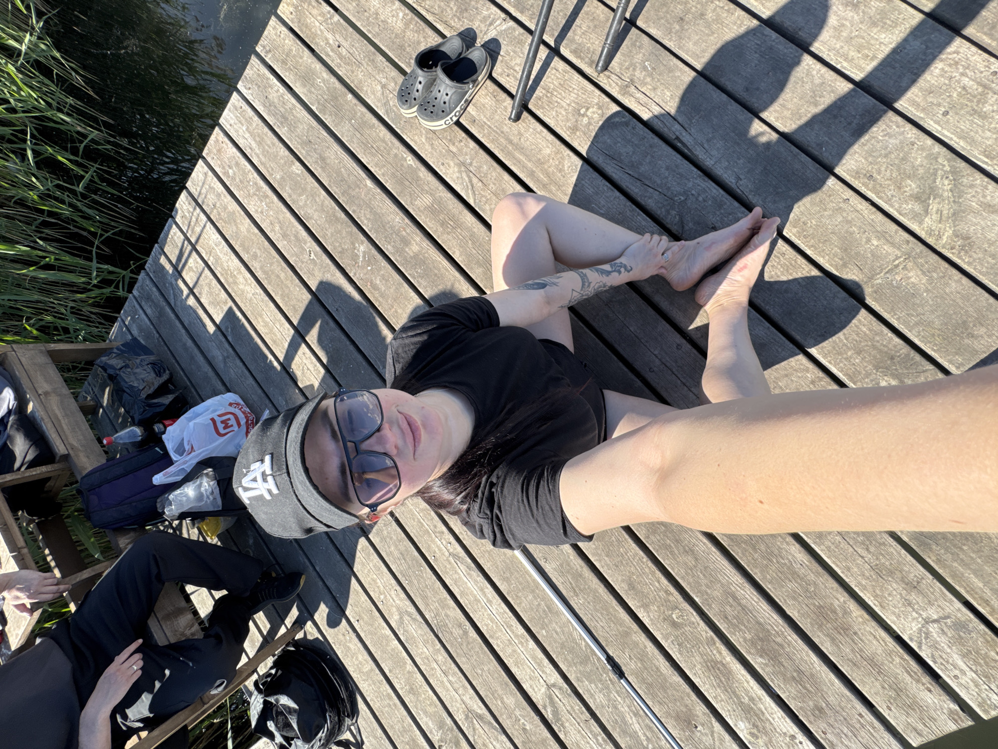 A high-angle shot depicts a person with dark hair, wearing sunglasses and a baseball cap with the letters "LA" on it, lying on a wooden dock. They are wearing a black t-shirt and shorts. Their bare feet are visible near the top right corner of the frame. A pair of gray Crocs and the legs of a metal chair are also in the upper part of the image. To the left of the person, a second person dressed in black is sitting, and there is a collection of bags, including a bag from a supermarket chain. The dock extends to the left, where reeds and dark water can be seen. The environment suggests a lakeside or riverbank setting, though the location is unknown. A fishing rod is also nearby. The image is brightly lit, with strong shadows indicating a sunny day.