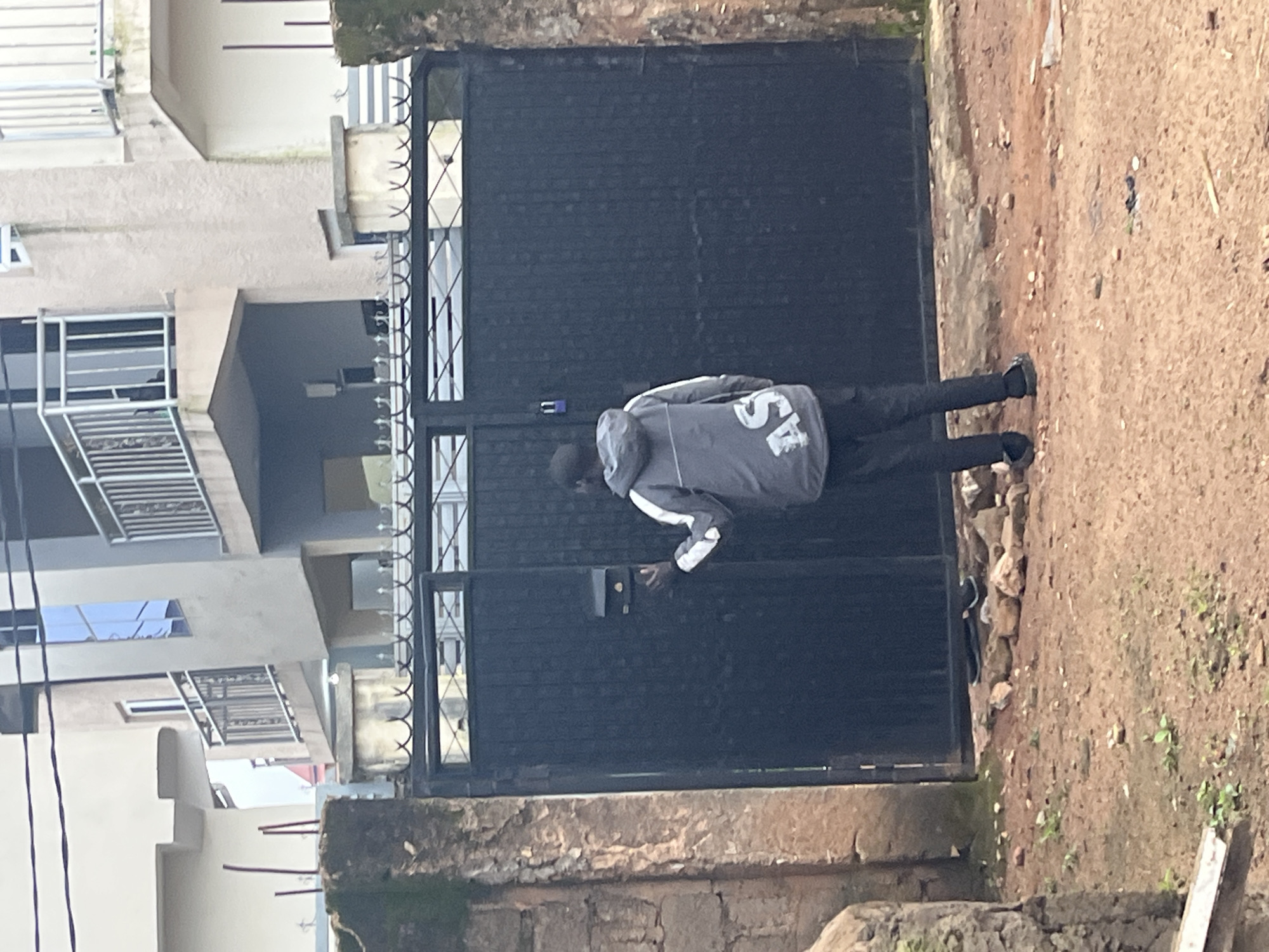 A full shot shows a person standing in front of a large, black, metal gate. The person, who appears to be male, is wearing a grey hooded jacket with white trim on the sleeves and "AS" printed in white on the back. He is also wearing black pants and black slip-on shoes. The person's right hand is extended towards the gate, and he appears to be interacting with a small, dark object mounted on the gate, possibly a mailbox or intercom.
The gate is tall and appears to be a single, solid panel. Above the gate is a decorative metal railing with sharp spikes. To the left of the gate is a section of a stone wall.
Behind the gate, partially visible, is a modern-looking building with balconies and large windows. The building is light-colored with grey accents.
The ground in front of the gate is covered in red earth, with scattered stones and a few small green plants. The overall scene suggests an outdoor environment, possibly a residential or commercial property. The lighting indicates it is daytime, but overcast. The image was taken in Jos, Nigeria.