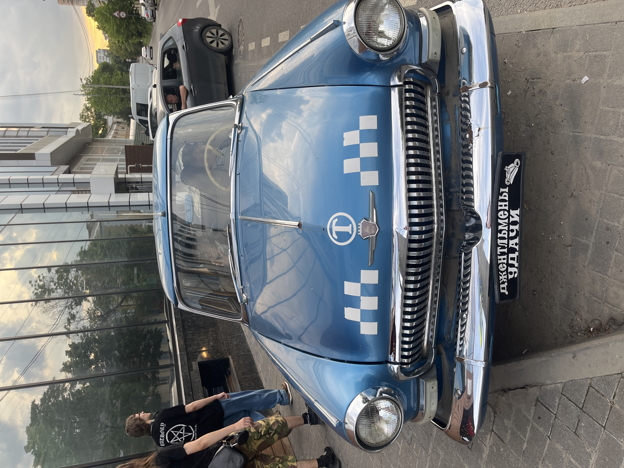 A blue vintage sedan, configured as a taxi, is prominently featured on a city street in Krasnodar, Russia, during what appears to be late afternoon or early evening. The vehicle, likely a GAZ-21 Volga, is painted a bright blue with a chrome grille, circular headlights, and a chrome front bumper, which exhibits visible dents and scrapes. The hood displays a white "T" in a circle emblem and a white checkered pattern, signifying its taxi function. A black decorative license plate below the grille bears the Cyrillic text "ДЖЕНТЛЬМЕНЫ УДАЧИ" (Gentlemen of Fortune). A driver is discernable within the vehicle through the windshield.
On the paved sidewalk to the left of the car, two individuals are partially visible; one wears a black t-shirt featuring a white pentagram and the word "BATHORY," while the other wears camouflage shorts and carries a black shoulder bag. Adjacent to the blue sedan, in the street, a grey hatchback is stopped, with its male driver and a younger passenger visible through its open windows.
The immediate surroundings include a multi-story modern building with a reflective glass facade on the left, mirroring portions of the sky and trees. The sidewalk is paved with grey rectangular blocks. The street is asphalt, separated from the sidewalk by a curb. In the background, additional urban buildings, green foliage, and other vehicles are present, along with a "40" speed limit sign. The sky in the upper right portion of the image shows warm, orange light, consistent with sunset.