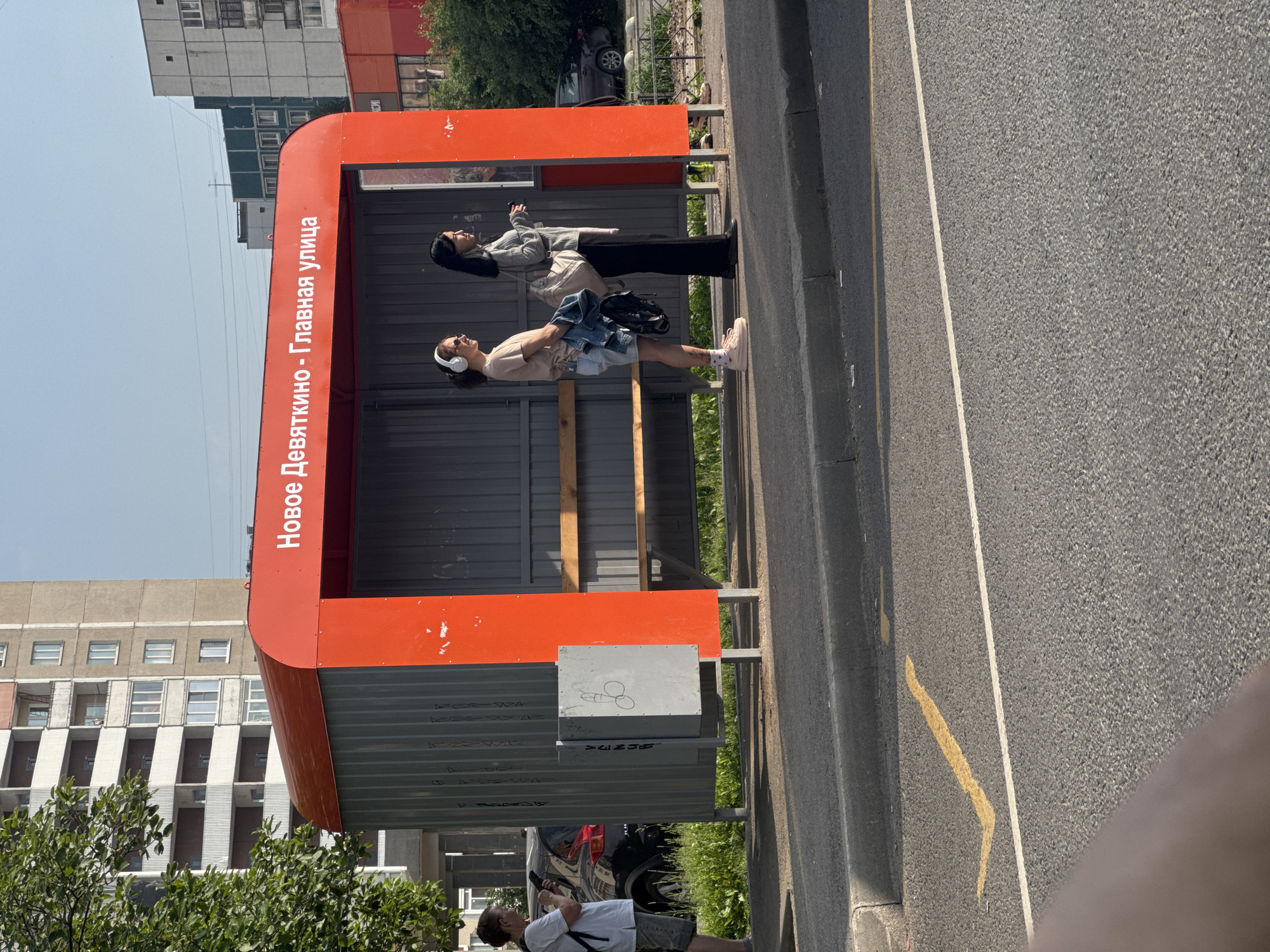 Two women stand at a bus stop in Novoe Devyatkino, Russia. The bus stop has a red roof and gray metal walls, with the words "Новое Девяткино - Главная улица" (Novoe Devyatkino - Main Street) written in white on the roof. One woman on the left wears headphones and a light brown t-shirt with denim shorts, holding a denim jacket. The woman on the right wears a gray sweater and black pants, carrying a large beige bag. A silver garbage bin with graffiti is located to the left of the women. In the background, a modern residential building and trees are visible. The bus stop is adjacent to a paved road with yellow road markings.