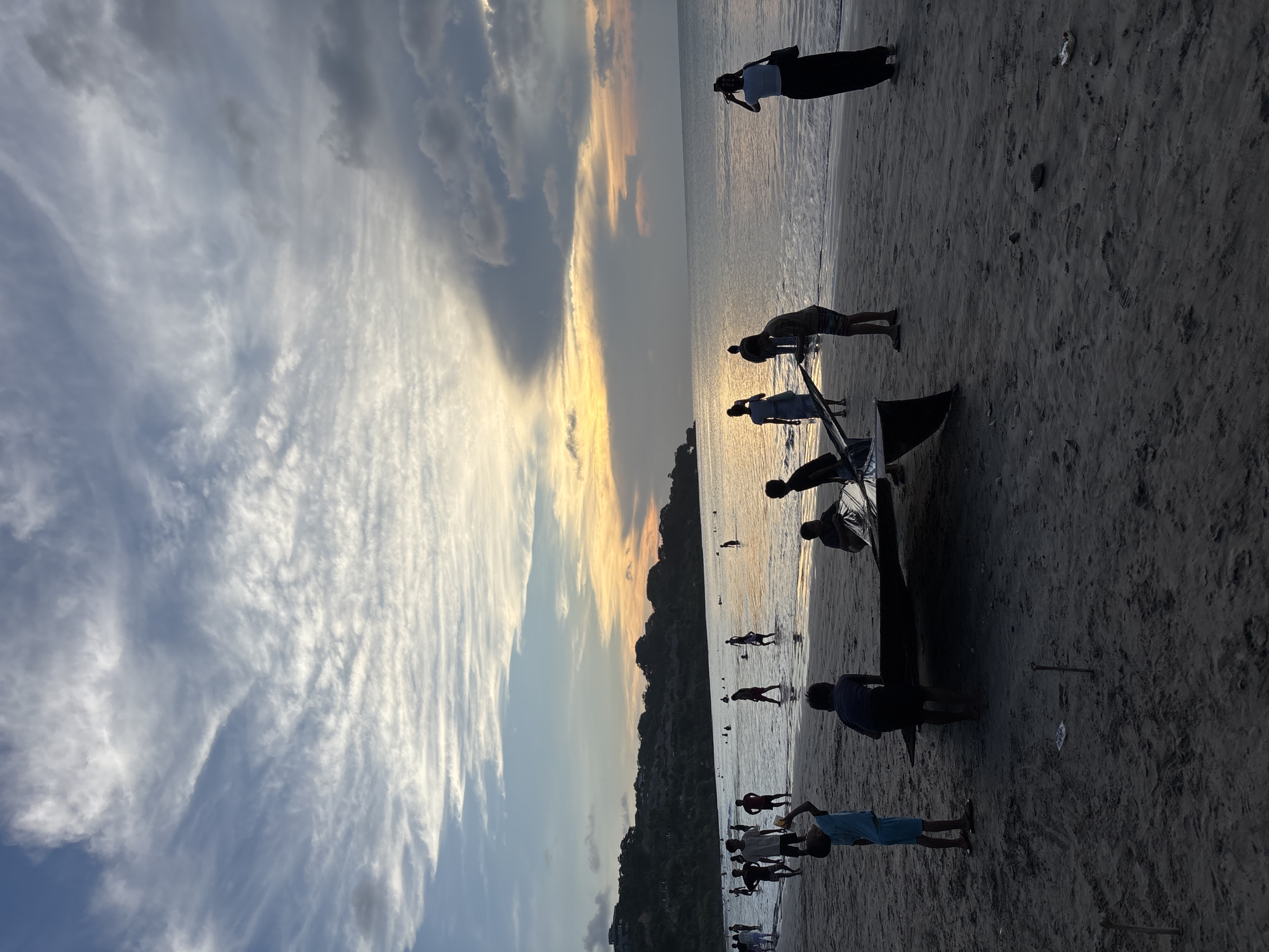 The image shows a beach scene at sunset in Kuta Selatan, Indonesia. The sky is filled with striated clouds, transitioning from blue-gray to golden hues near the horizon. A dark landmass is visible along the horizon line. Silhouetted figures of people populate the beach. Some are near a dark boat that is positioned on the sand. Others appear to be wading or standing in the shallow water. There is a distinct tide line and a moderately textured sand surface.