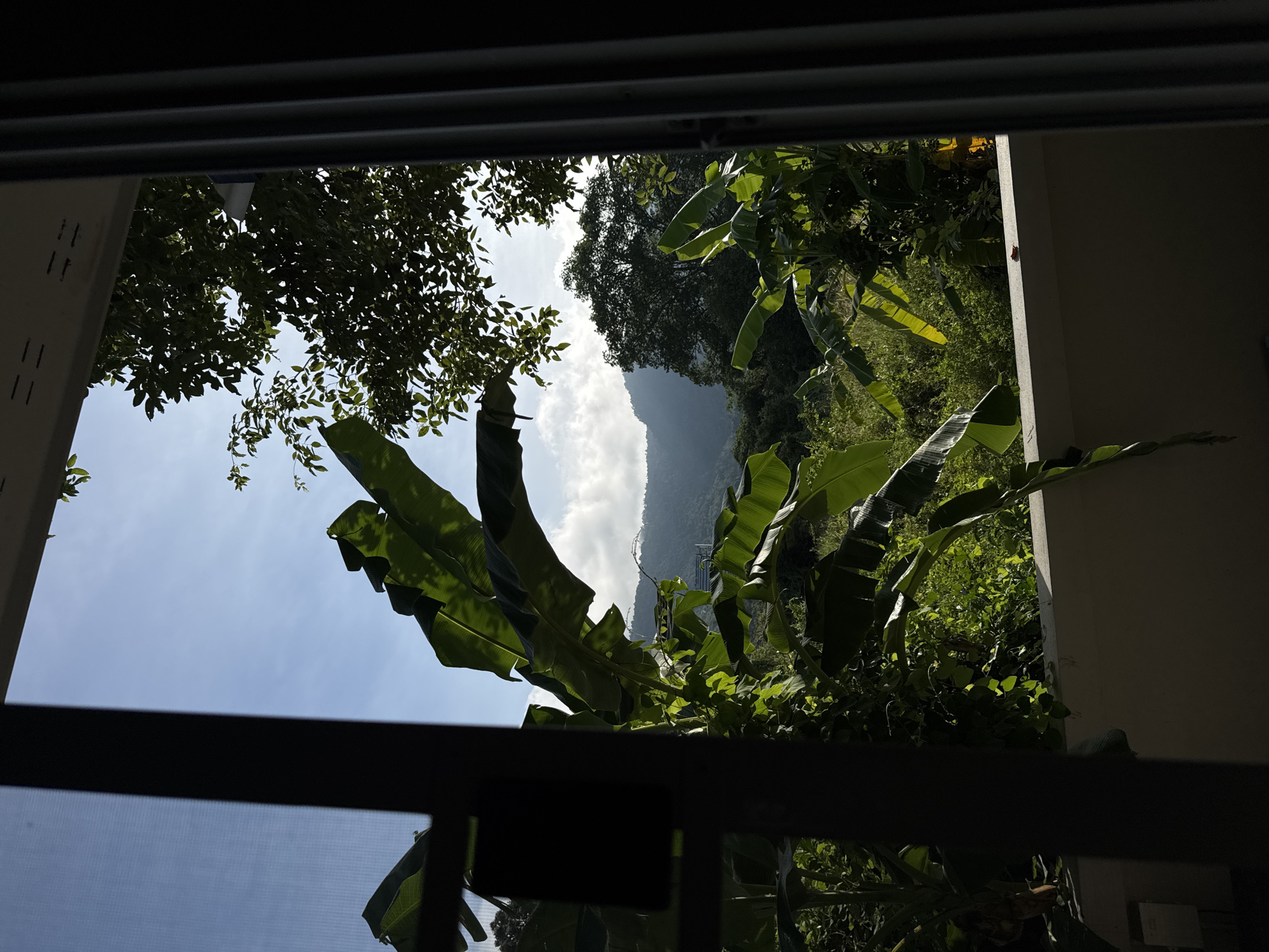A view through a window or doorway shows a lush, green tropical landscape with mountains in the distance. The foreground is dominated by large, dark green banana leaves, some of which are lit by sunlight. A dark, vertical post is present on the left side of the frame, obscuring a portion of the scene. The middle ground features more dense vegetation and a large tree. The background consists of rolling hills and mountains, with some clouds in the bright blue sky. The scene appears to be from Baan Nok Sai, Thailand.