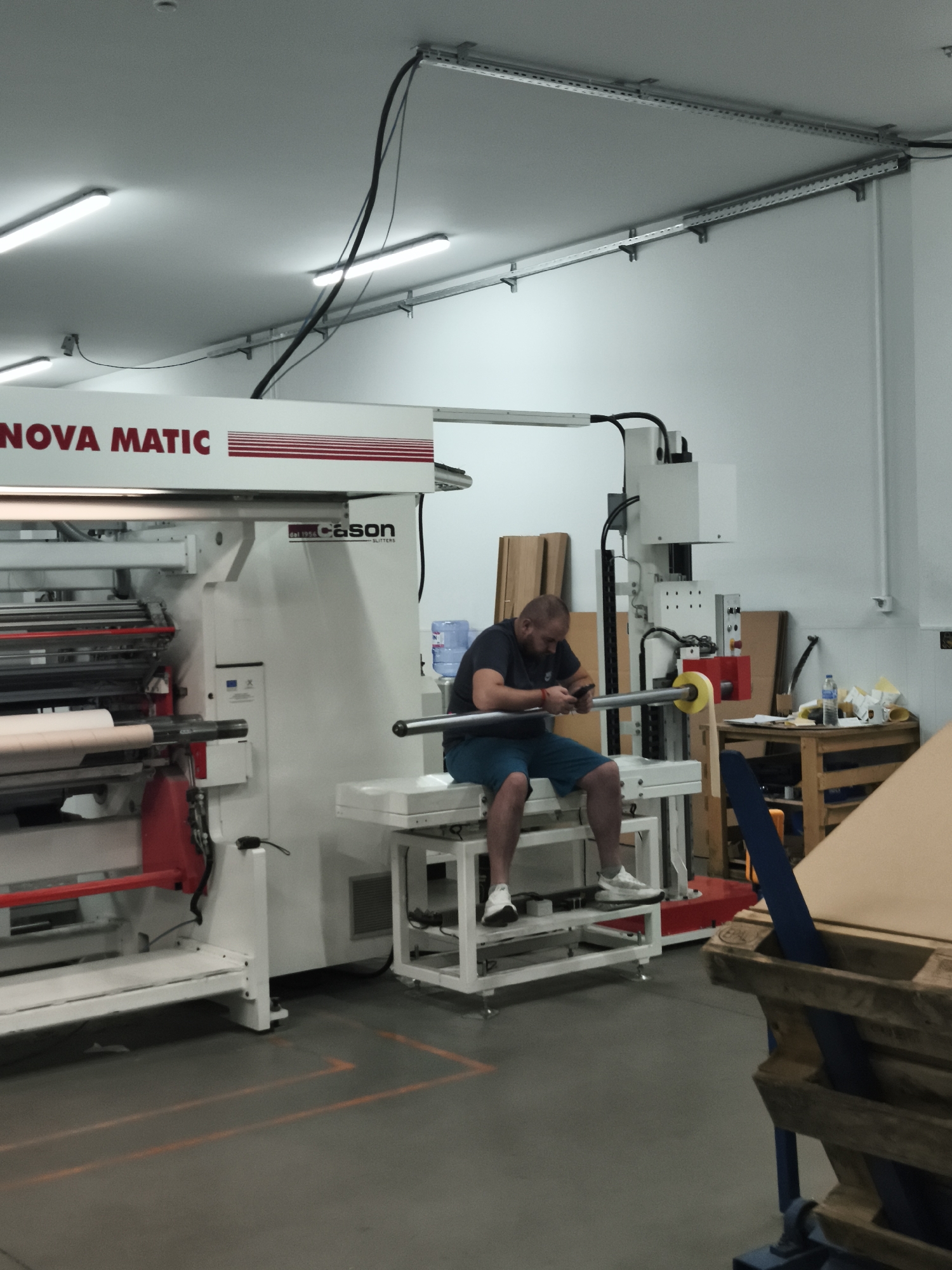In an industrial interior, a bald man in a dark t-shirt and blue shorts sits on a white platform, looking at his phone. He is holding a long, cylindrical metal pole with a yellow disk at the end. The man is positioned in front of a large, white and red industrial machine with the "NOVA MATIC" logo visible on its side. The machine appears to be a slitter, as indicated by the "CASON SLITTERS" text on its casing. In the background, there are cardboard boxes, a wooden table with various items, and a water cooler. The floor is grey concrete with orange painted lines. Overhead, fluorescent lights illuminate the space, and industrial conduits run along the ceiling.