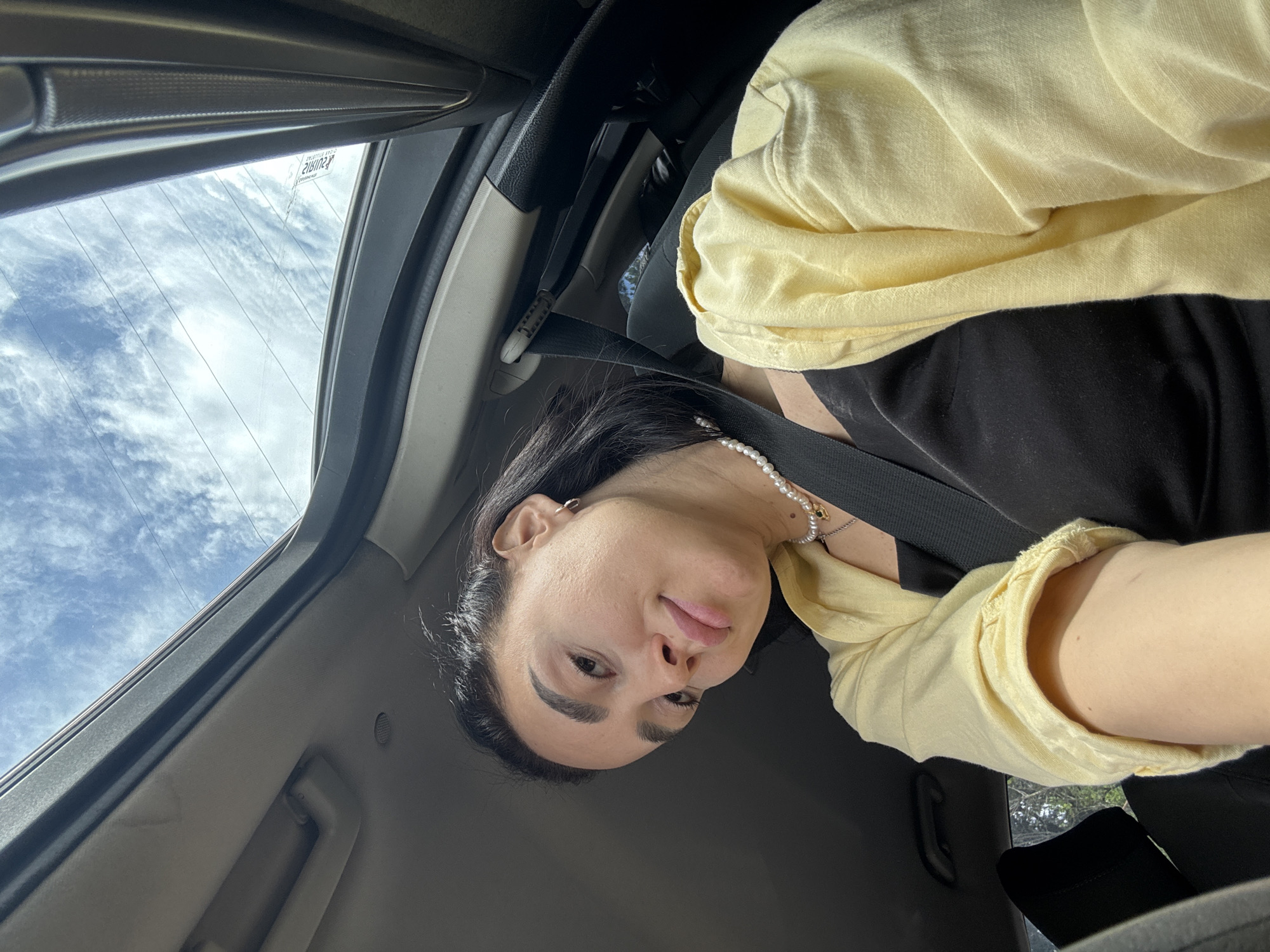 A woman is sitting in the driver's seat of a car, wearing a black top, a yellow shirt, and a pearl necklace. She is wearing a seatbelt and appears to be taking a selfie. The sky with clouds is visible through the car's window. There is no information to determine if this image was taken in Tbilisi, Georgia.