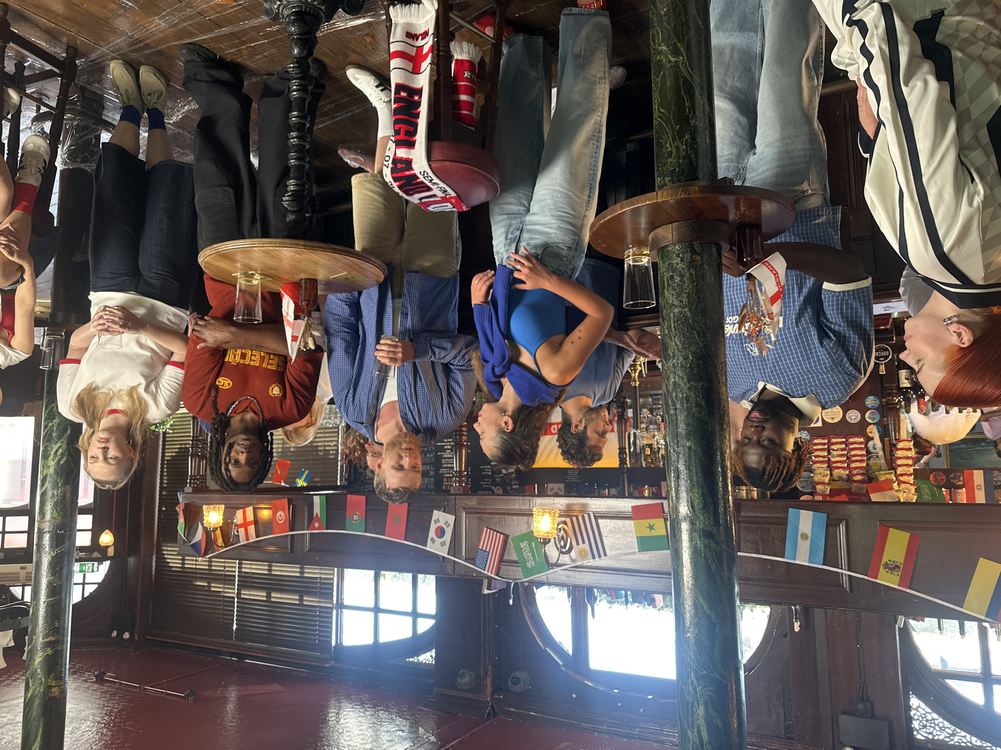 This appears to be a social gathering inside a pub. The image is oriented upside down. Several individuals are standing around tables, engaging in conversation and holding drinks. The bar is decorated with various national flags. An "England" scarf is visible near a table leg. Based on the "DORCH" label visible and the England details, it is probable that the location is the Dorchester in London, United Kingdom, an establishment known for its links to England football.