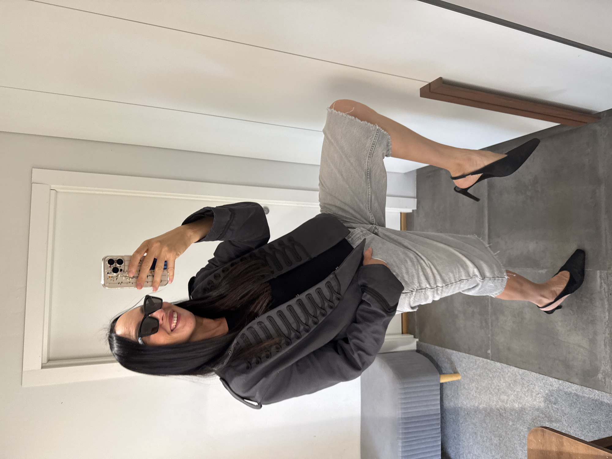 A person is standing in a room and taking a selfie in a mirror. The person is wearing a dark grey military-style jacket with braiding, a black t-shirt, light grey denim shorts with a raw hem, and black slingback heels. The person is also wearing sunglasses and has long dark hair. The room appears to be a home or fitting room, with a white door, a grey upholstered bench, a grey tiled floor, and a partial view of a wooden object. There is no indication that the image was taken in Chaikovsky, Russia.