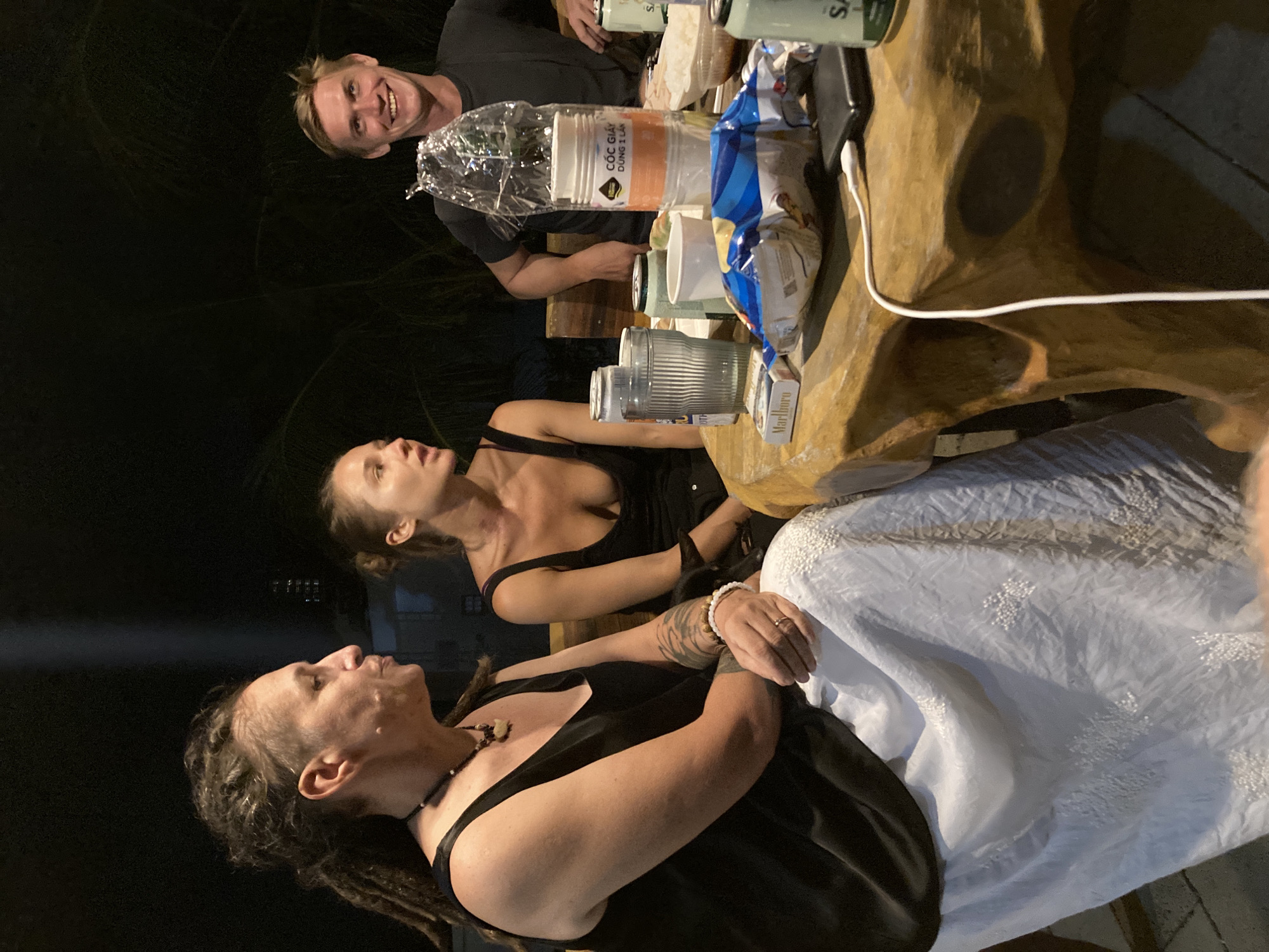 A group of people is gathered around a table at night. There are three people visible: a man with fair skin and short blond hair is at the top of the frame. Two women are below him, the first with long dark hair pulled back into a ponytail and the second with dark dreadlocks. They are all seated at a wooden table with drinks, cups and other objects. The location could be in Mũi Né, Vietnam, given the writing on the plastic wrap is Vietnamese.
