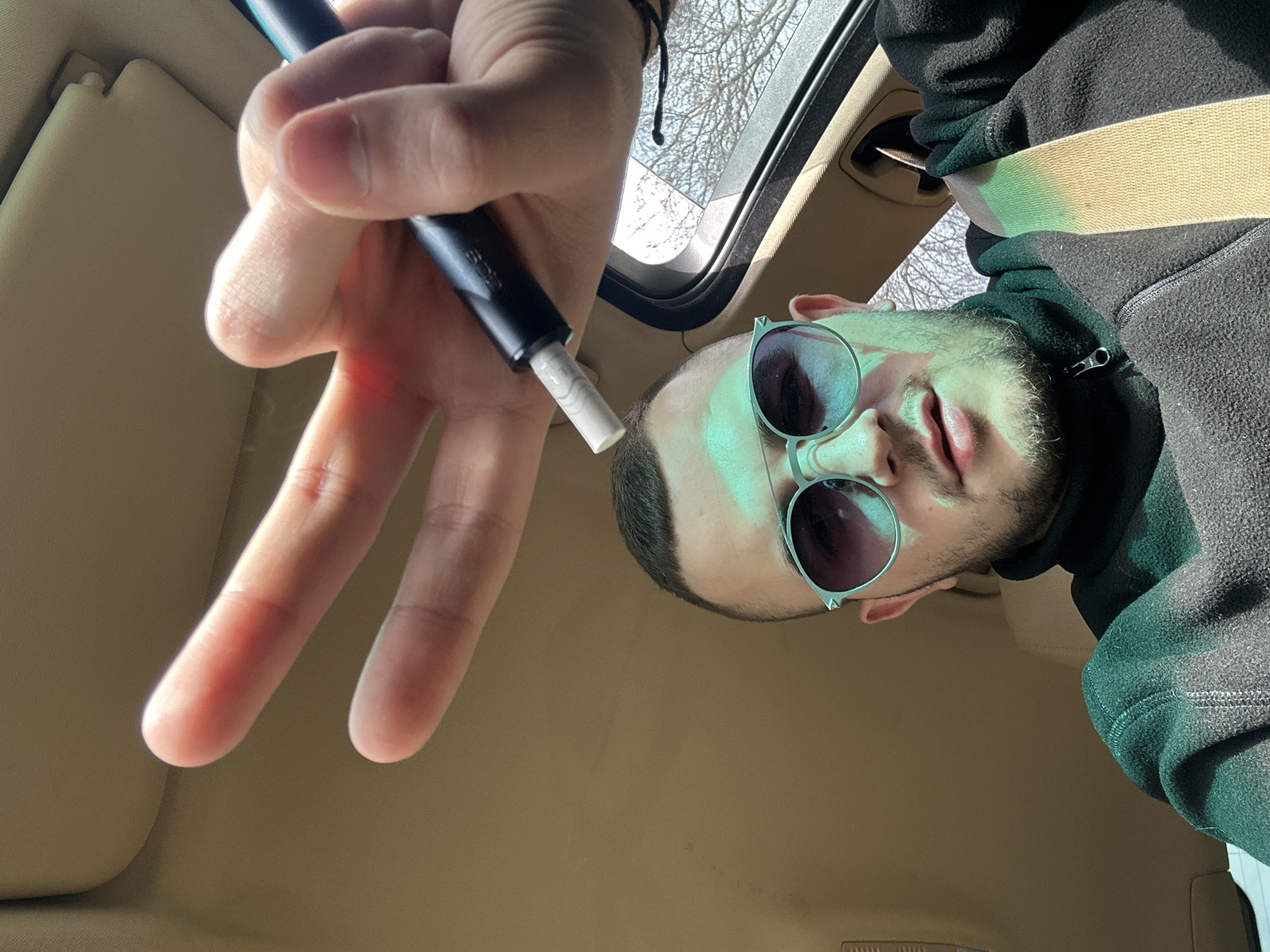 A man is inside a vehicle with a beige interior. He is wearing sunglasses and a dark-colored hoodie. A hand holding a vape pen with a white filter is in the foreground. The background shows part of the car's sunroof and trees. The location is unknown; there is no definitive information to identify Plovdiv, Bulgaria.