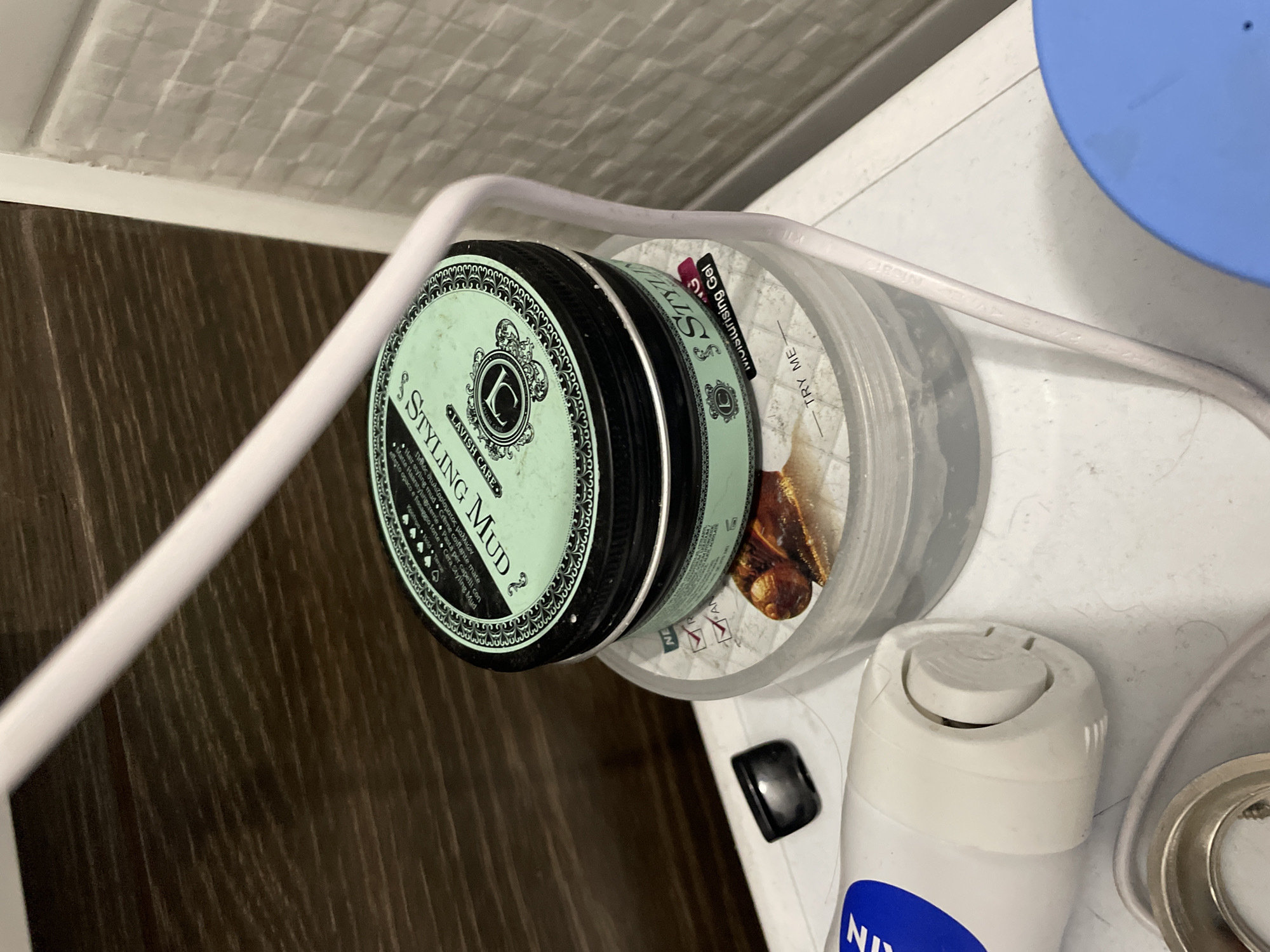 The image depicts a close-up, high-angle shot of several personal care items on a white surface, possibly a countertop or shelf, in what appears to be a bathroom. The background features a dark wood-grain texture on the left and a tiled wall on the right.
Dominating the center of the frame is a mint-green jar with a black lid, labeled "STYLING MUD." The lid has intricate black decorative elements and a stylized "L" logo. This jar is stacked on top of a larger, clear plastic container. The clear container has some text and imagery visible, including the words "TRY ME" and a portion of a label that appears to say "Styling Gel."
To the left of the main stack is the cap end of a white tube with a blue stripe. The cap has a visible dispensing mechanism. Below this tube, a metallic ring is partially visible.
A thin, light-colored wire, possibly from an electronic device, arcs across the middle and right side of the image, passing behind the styling mud jar. In the lower right corner, a bright blue lid of another container is visible.
The lighting is bright, casting some shadows. The overall scene suggests a collection of grooming or cosmetic products. There are no people visible in the image. The environment appears to be indoors, with the textures suggesting common bathroom or vanity finishes. The location is Sinyavino, Russia.