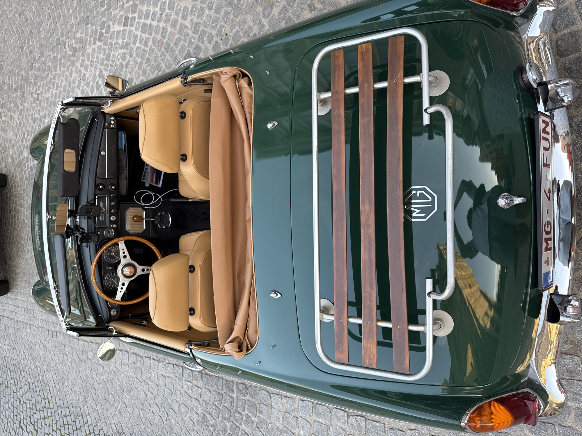 This media file shows the interior and rear of a vintage dark green MG convertible, parked on a cobblestone street. The car has tan leather seats and a tan convertible top that is partially down. The dashboard is visible, with a wooden steering wheel and multiple gauges. A luggage rack with wooden slats is mounted on the trunk.
The scene appears to be outdoors during the daytime, with the sun casting reflections on the car's polished surface. The cobblestone street suggests an older, European setting. The image does not show any people or indicate a specific event taking place.
Notable details include the chrome trim on the car, the "MG" logo on the trunk, and the license plate which reads "MG - 4 - FUN". The reflections on the car suggest the presence of buildings, possibly historic architecture, consistent with a European city. The overall impression is one of classic automotive beauty in a charming historical location. The context provided identifies the location as Bruges, Belgium.