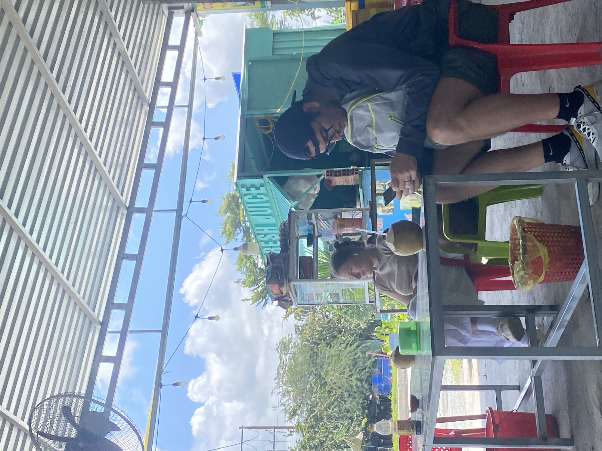 A man and a woman are sitting at a table outside a food stall in Ben Cau, Vietnam. The stall has a sign that says "FRESH JUICE" and is painted green. The man is wearing a blue baseball cap, glasses, and a dark jacket. He is looking down at his phone and has a grey backpack on his lap. The woman is wearing a light brown shirt and is looking to her left. There are two coconuts and a green cup on the table. In the background, there are trees and a blue sky with clouds. A large fan is mounted on the ceiling above the table.