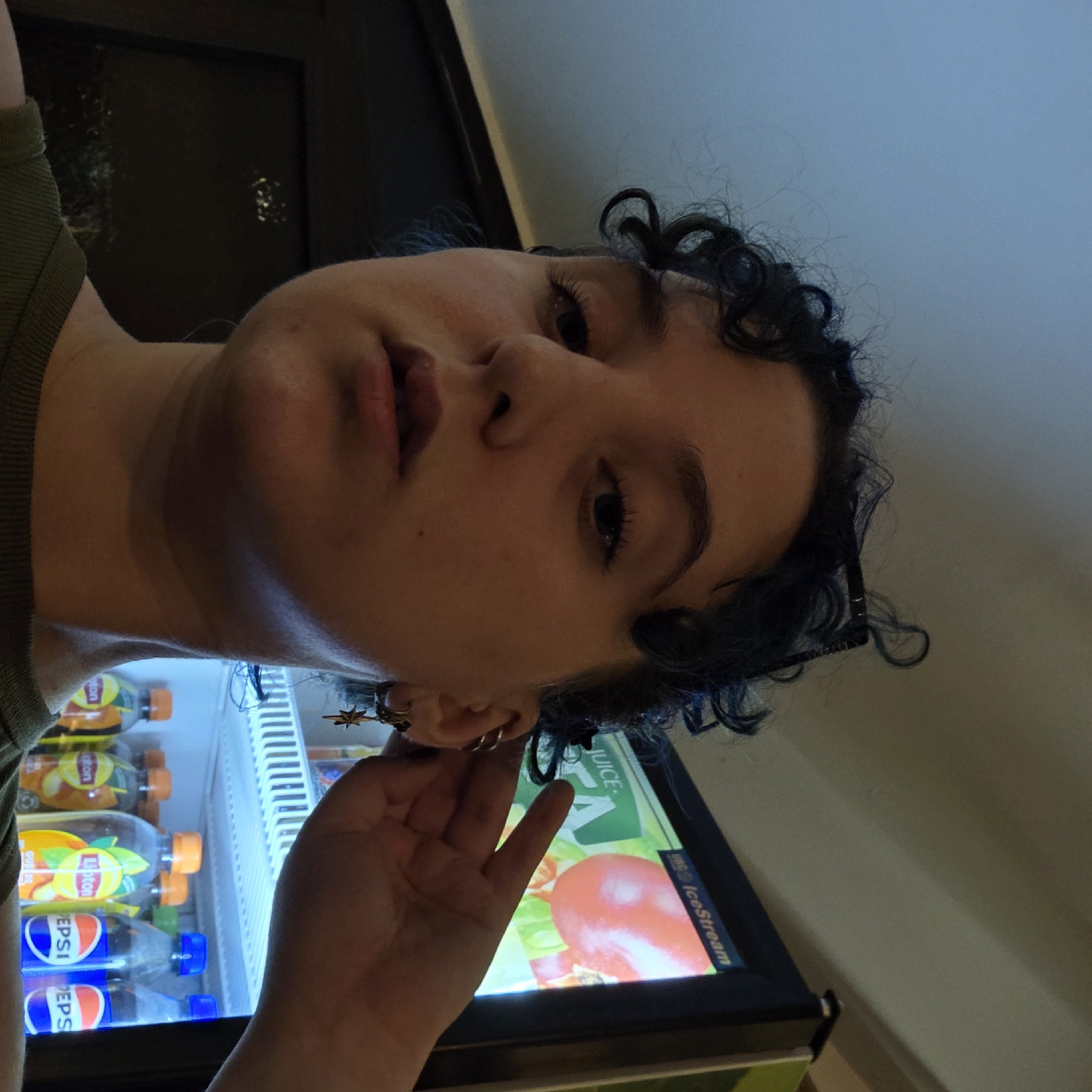 A close-up selfie shows a person with curly dark hair, some of which is dyed blue. The person is looking towards the camera with their right hand touching their ear, where they are wearing multiple earrings, including one with a star charm. Behind the person, a brightly lit cooler or refrigerator is visible, stocked with bottles of Lipton iced tea and Pepsi. The cooler has a screen displaying a graphic of a red tomato and the words "JUICE" and "IceStream". The lighting in the scene is artificial and cool-toned, creating a somewhat dim atmosphere. The background suggests an indoor setting, possibly a store or a public place with vending machines. The image does not provide any specific clues to identify the location within Chișinău, Moldova.