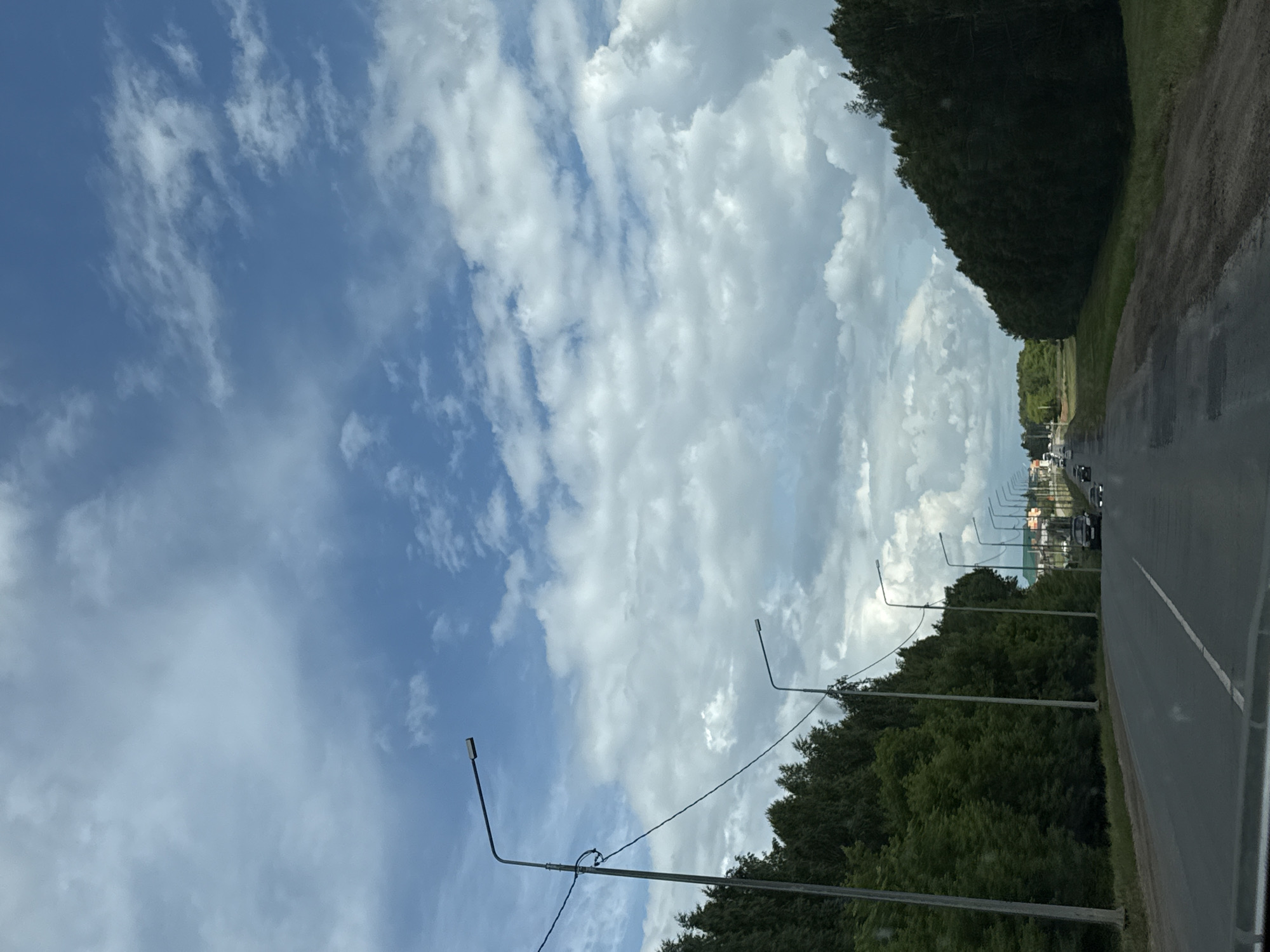 The image depicts a two-lane asphalt road winding through a tree-lined landscape. The road has a white dashed line marking the center. Several streetlights are visible along the left side of the road. The environment appears to be a rural or suburban area, with dense green trees lining the road on both sides, particularly on the left. In the distance, there are buildings and more trees. Several cars are driving on the road. The sky is filled with dramatic clouds, suggesting a dynamic weather system. The perspective is from inside a vehicle, looking forward along the road. There are no people directly visible in the image, only the presence of vehicles on the road. The overall scene suggests a journey or travel through a picturesque, natural setting.