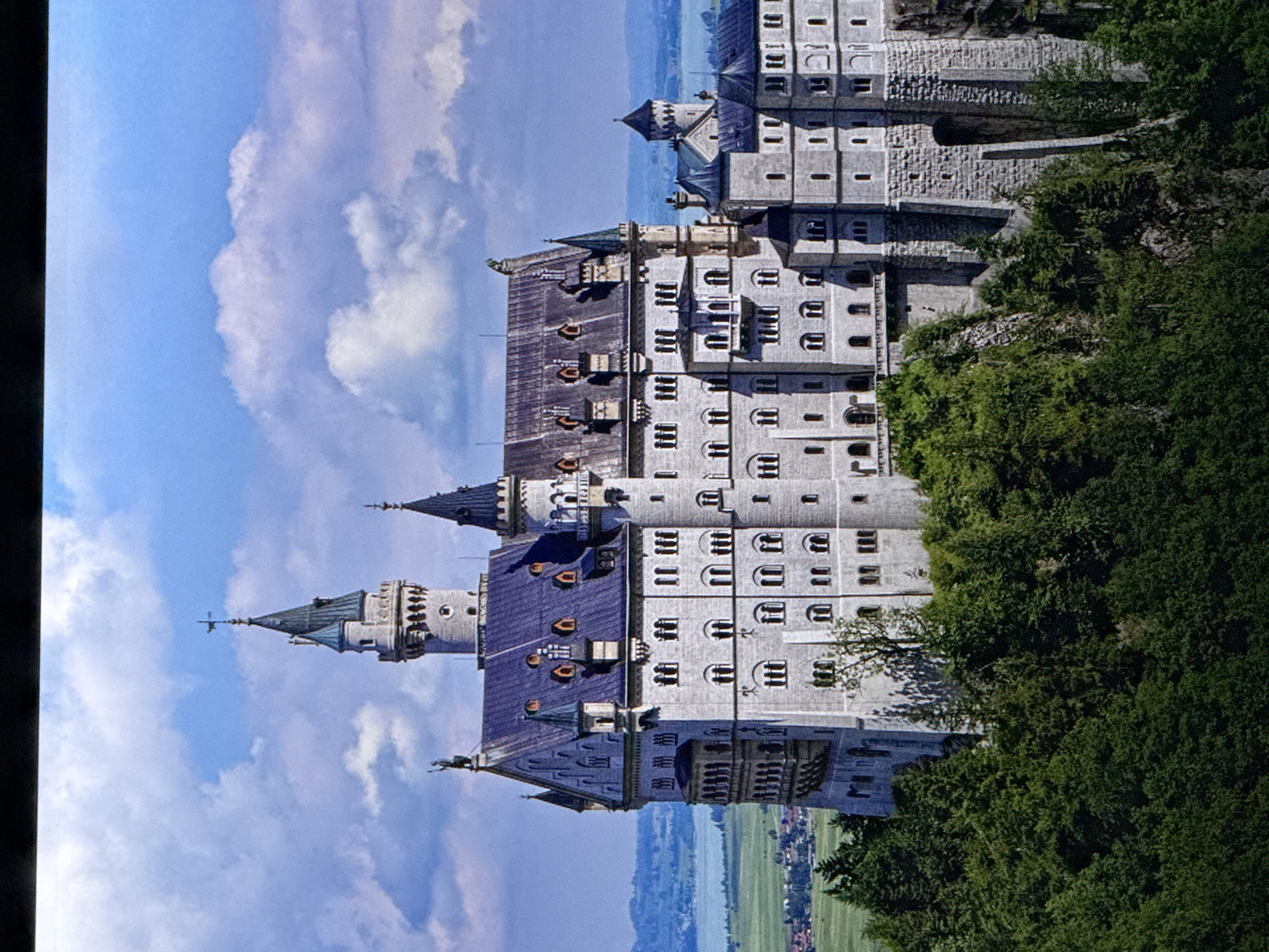 The image shows a grand, fairytale-like castle perched on a verdant hillside, with a body of water and rolling green hills in the background under a bright blue sky with scattered white clouds. The castle itself is a complex structure with multiple towers, turrets, and steeply pitched roofs, constructed of light-colored stone. It features numerous windows and architectural details like balconies and decorative stonework. The surrounding landscape is characterized by lush green trees, particularly in the foreground, and a gently sloping terrain leading down to a more cultivated area with buildings and fields. The overall impression is one of majesty and picturesque scenery. This castle is Neuschwanstein Castle, located in Bavaria, Germany, not in Saratov, Russia.