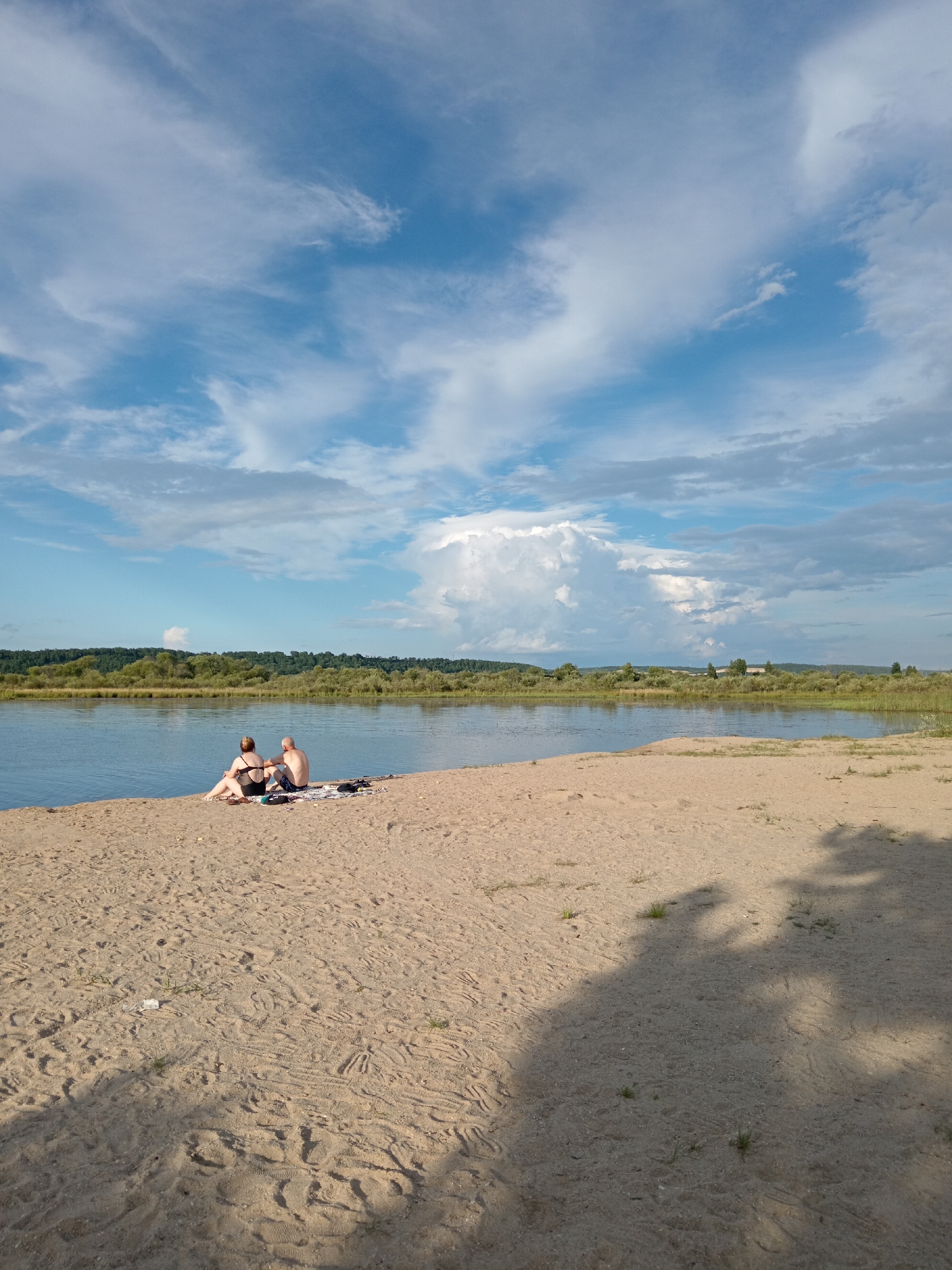 A couple sits on a sandy beach facing a body of water. The man is on the right, wearing blue swim trunks. The woman is on the left, wearing a black bikini. They are sitting on a patterned towel. In the distance, there is a lush green treeline and a cloudy blue sky. The scene appears to be a serene recreational area, potentially near Belogorsk, Russia, given the natural landscape and common beach activities.