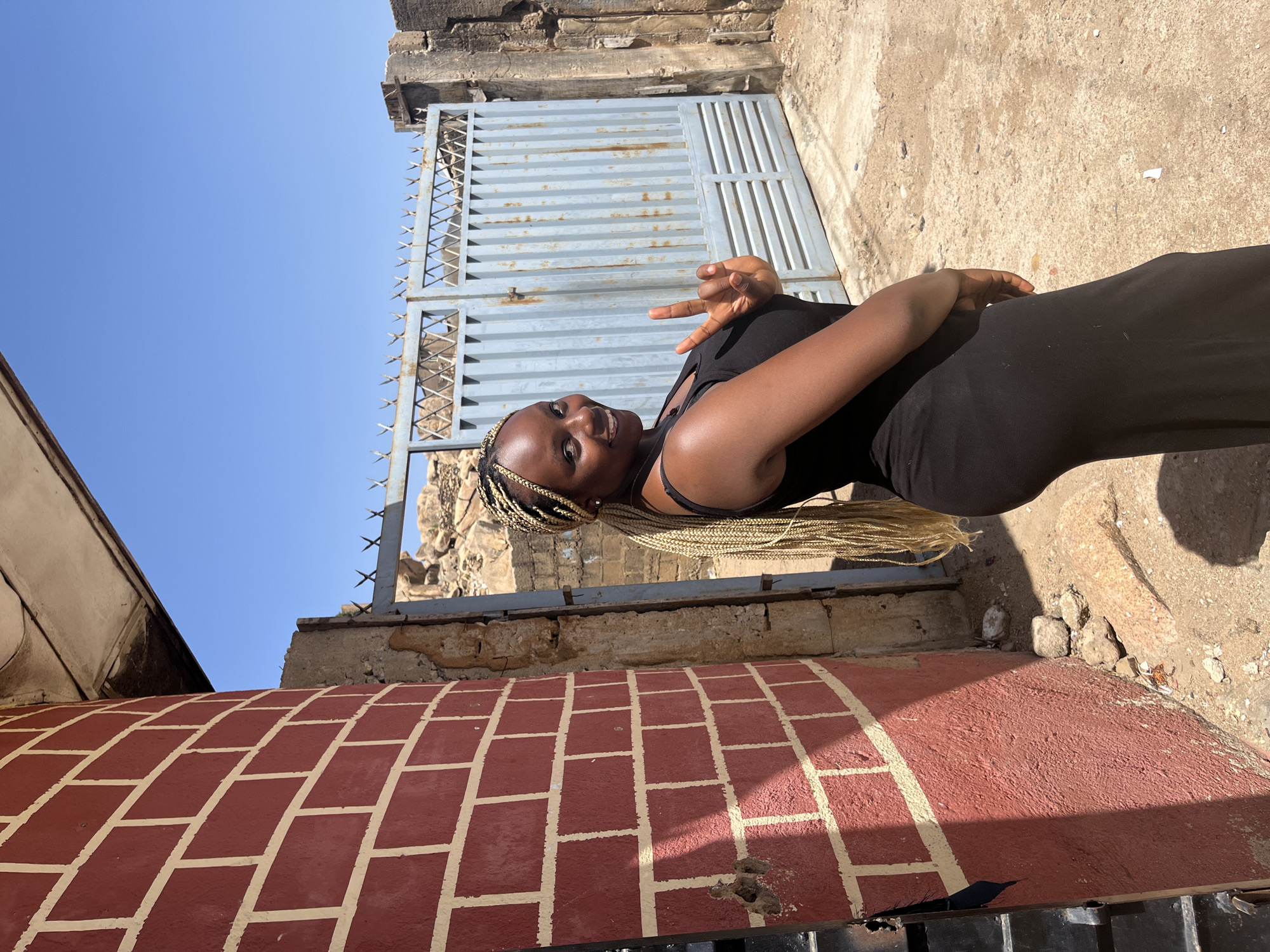 The image depicts a young woman in a black tank top and black pants posing outdoors. She has blonde braided hair and is holding up a peace sign with her left hand. Behind her is a weathered grey metal gate with spiked tops. The surrounding structure consists of brickwork, with some sections painted red with white lines. The background includes a clear blue sky. The location is a city of Unknown, Unknown.