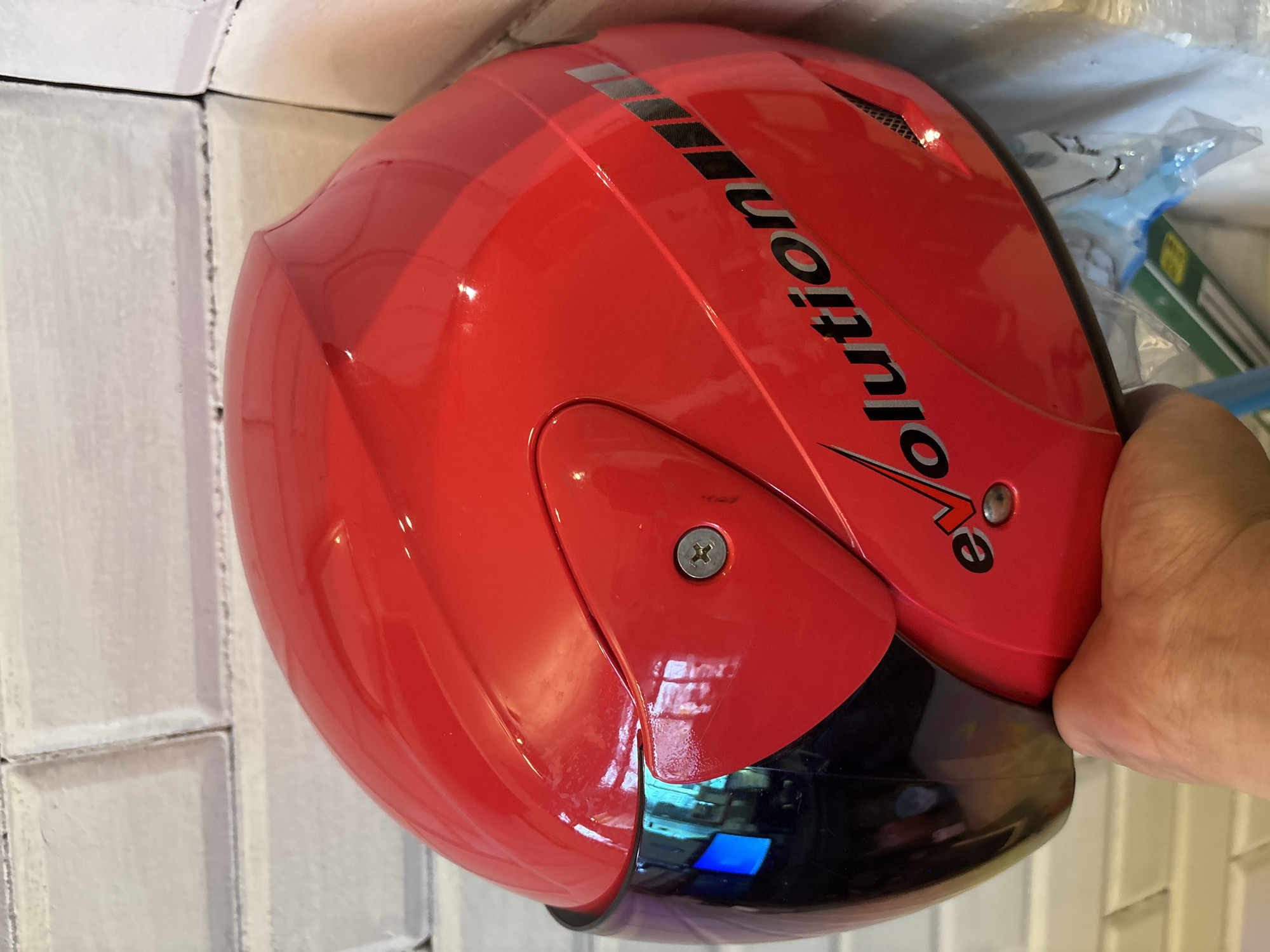 A red motorcycle helmet with a reflective blue visor is being held by a person's hand. The helmet has the word "evolution" printed on its side in black lettering with silver accents. The background appears to be a white tiled wall. The scene is a close-up shot, with the helmet dominating the frame.