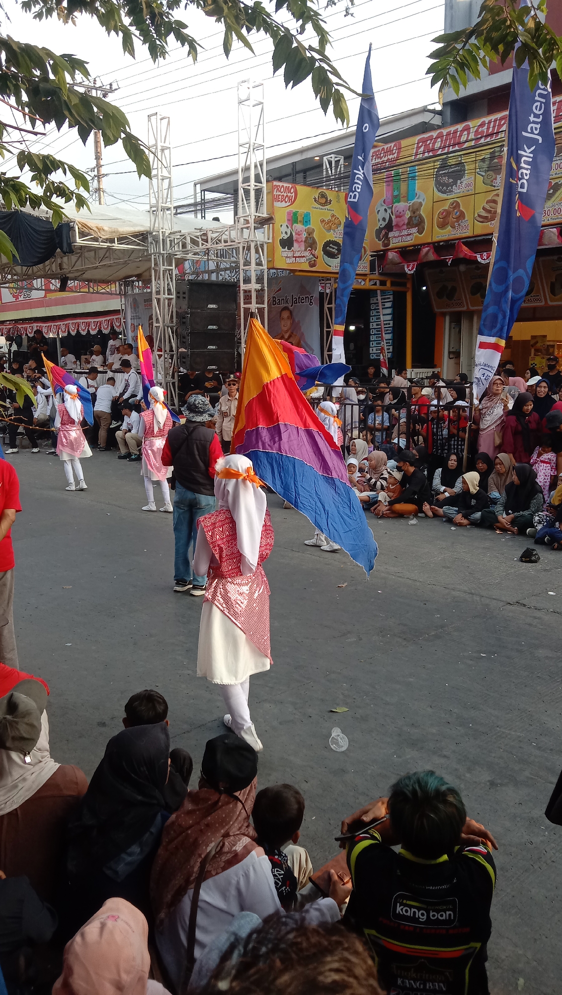 In Jatisawit, Indonesia, a street festival is underway, featuring a parade with performers in traditional attire. In the foreground, a young female performer, dressed in a white and red sequined outfit with a white headscarf, holds a large, multi-colored flag. Behind her, another performer in a similar outfit carries a smaller flag. Several other performers in matching costumes are visible further down the street, also holding flags. A crowd of spectators is gathered on the side of the street, sitting on the ground and watching the procession. Some onlookers are also standing. The background reveals a street lined with buildings, some adorned with banners and advertisements, including those for "Bank Jateng" and "Promo Heboh." A stage with a sound system is set up to one side. The atmosphere appears festive, with the parade and the gathering of people indicating a community event.