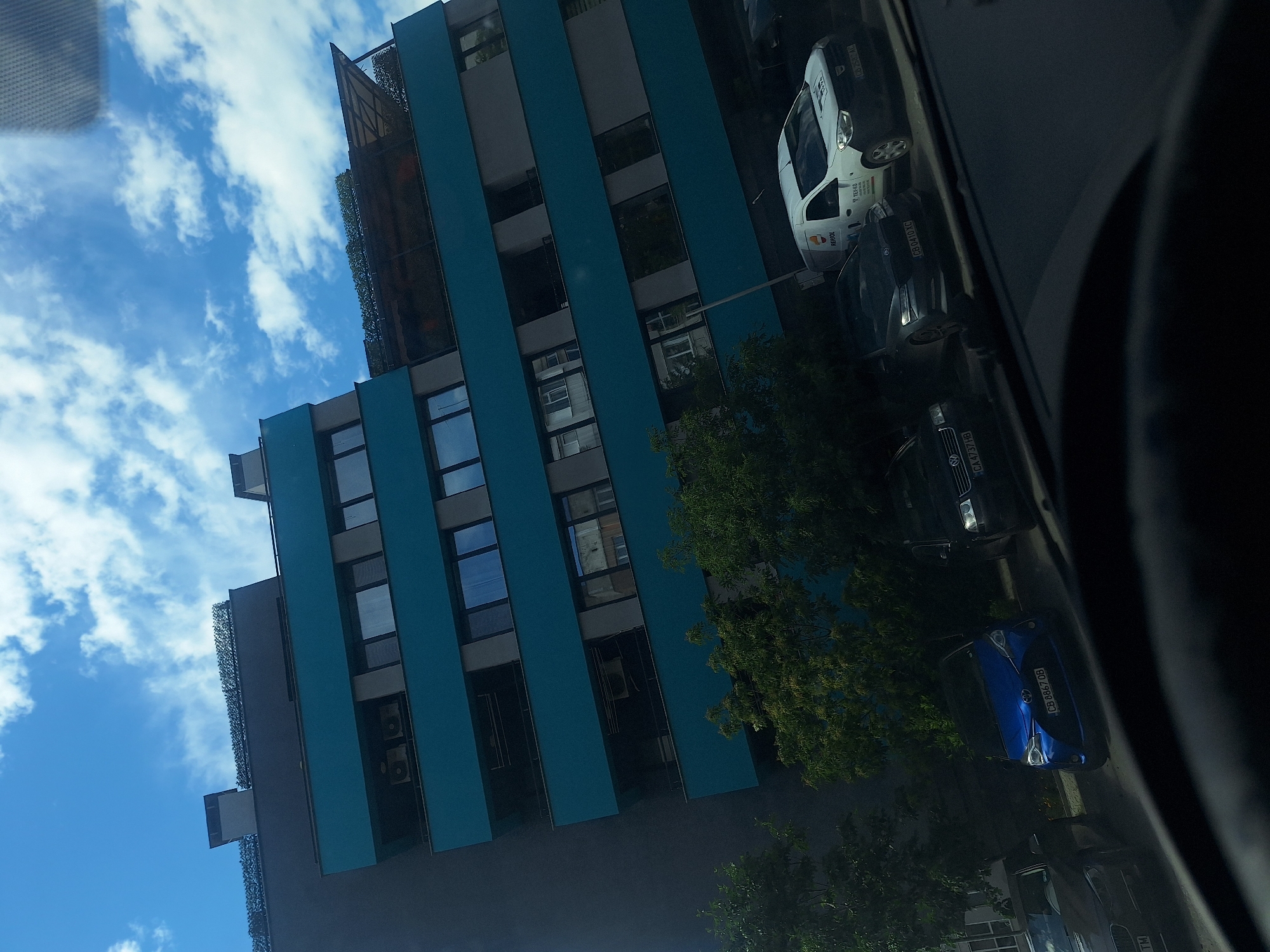 The image, taken from inside a car, captures a view of a multi-story building in Sofia, Bulgaria, under a clear blue sky with scattered clouds. The building has a modern design, characterized by alternating gray and turquoise vertical panels, with windows arranged in a grid-like pattern.
Several parked cars line the street beside the building. A white van with "REPJOL" written on its side and "TEX-KO" is visible. Other cars, including a blue one with the license plate "CB 8867 OB", a black one with the license plate "CA 4737 7HB" and another black one with the license plate "CB 0410 XC" are parked along the curb.
Trees with dense green foliage partially obscure the view of the cars and the building's lower sections. The interior of the car from which the photo was taken is visible on the right side, with elements of the dashboard and steering wheel in view.