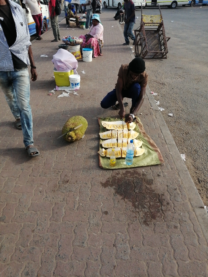 A man kneels, cutting a jackfruit laid on a green cloth on a sidewalk. He has already sliced part of the fruit into several pieces. Beside the sliced jackfruit is a bottle of water and a small jar. A whole jackfruit with a knife sticking out is nearby. People walk by in the background, and another woman sits with goods for sale. A cart stands nearby. The street is paved with a red brick pattern.