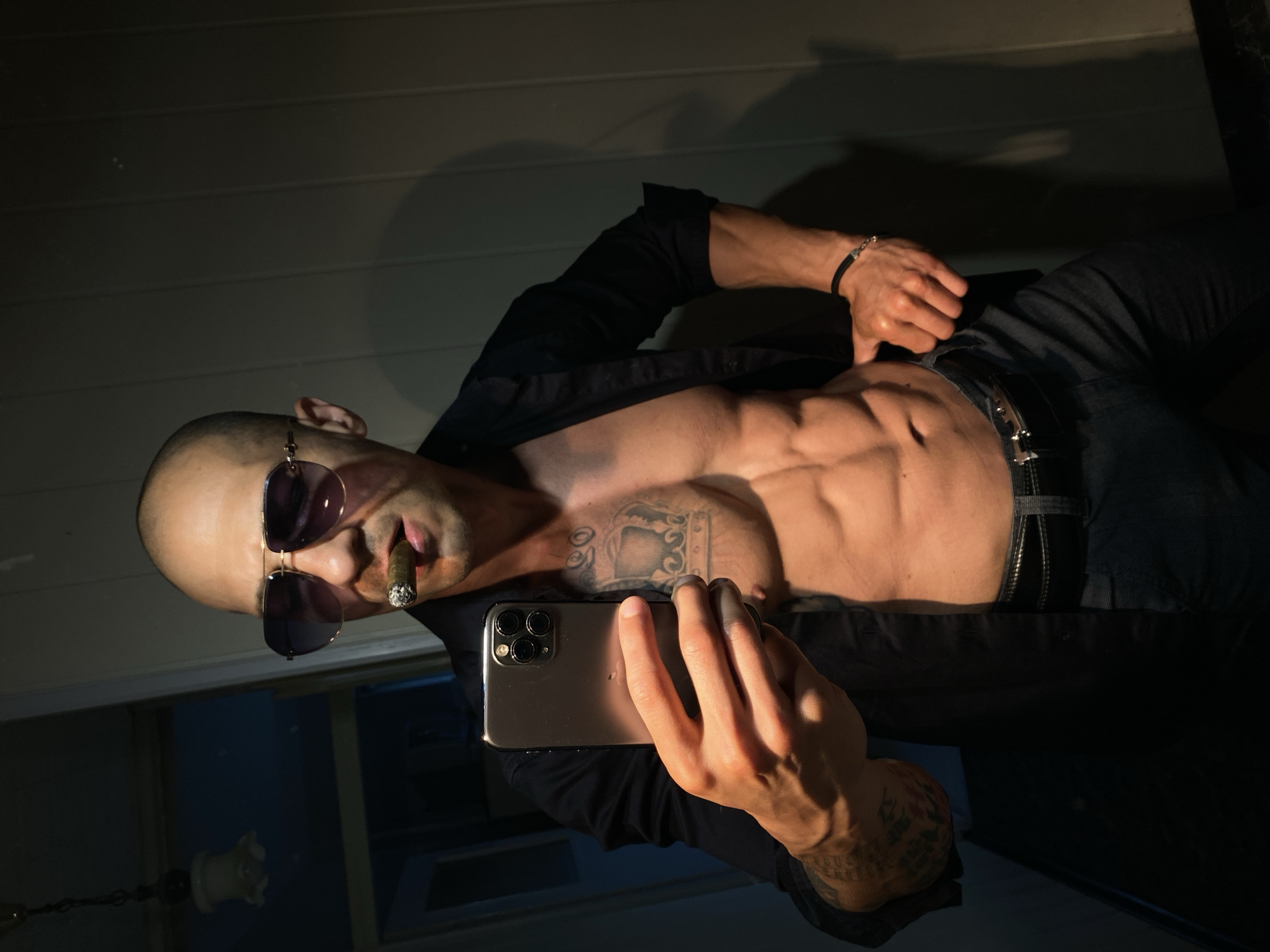 A man with a shaved head and sunglasses holds a smartphone, taking a selfie. He has a cigar in his mouth and is wearing an unbuttoned black shirt, revealing his bare chest and abdomen, which show well-defined abs and a chest tattoo. He is also wearing dark pants and a belt. The background appears to be an indoor setting with a wall and a dimly lit doorway. There are no recognizable landmarks or features that would suggest a specific location in Pleven, Bulgaria.