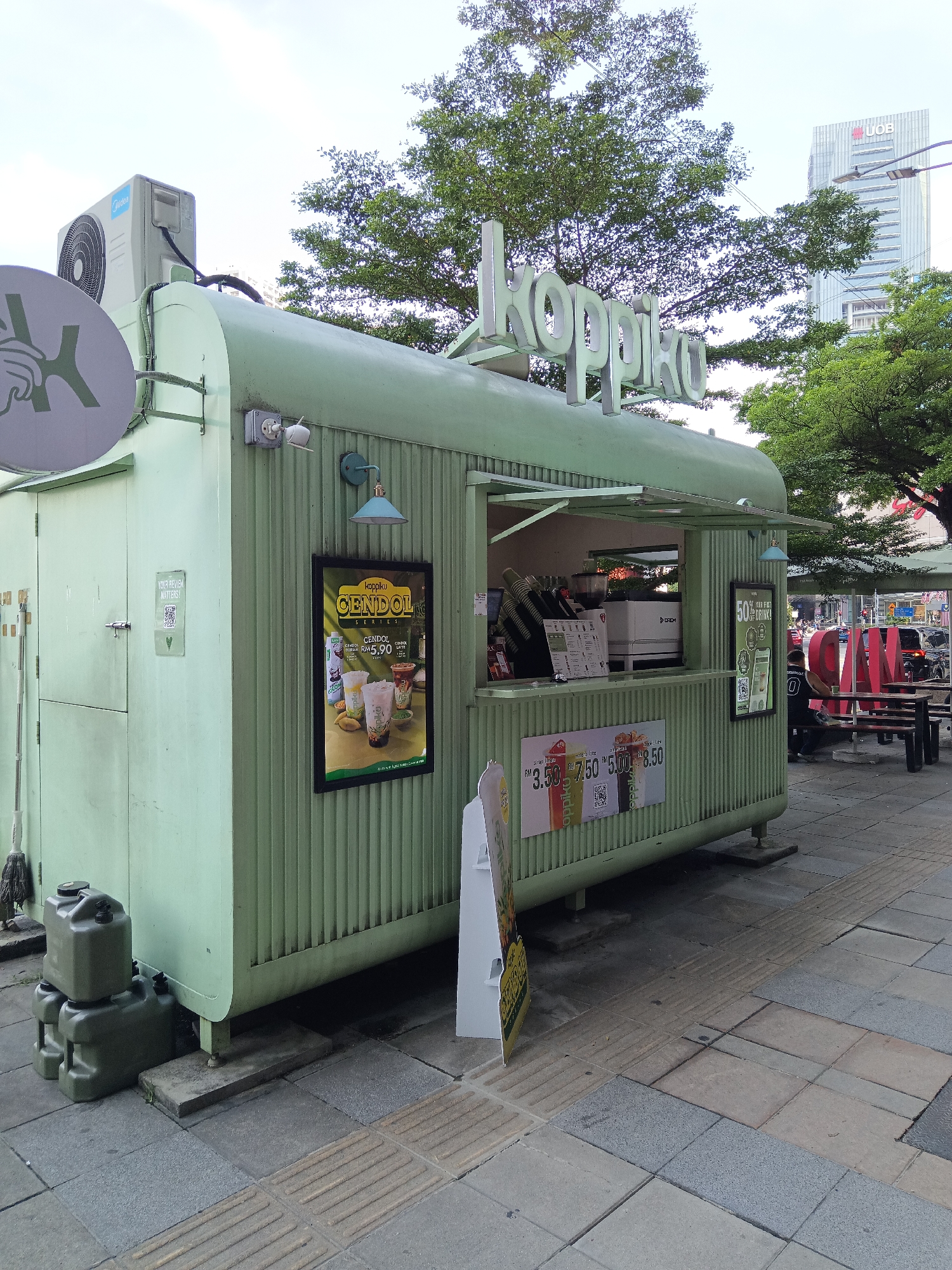 This is a medium shot of a light green, corrugated metal food kiosk in Kuala Lumpur, Malaysia. The kiosk has a serving window with a counter and an awning. Above the serving window, the white, three-dimensional letters "koppiko" are mounted. A large sign with the text "CENDOL" and prices in Malaysian Ringgit is displayed on the front of the kiosk. To the right of the serving window, a smaller sign advertises "50% DRINK!".
An air conditioning unit is mounted on the roof of the kiosk. To the left of the kiosk, there are several grey plastic water containers stacked on the ground. A broom leans against the side of the kiosk. In the background, trees and a tall building with the UOB logo are visible. Several people are standing and walking near the kiosk and in the background. The ground in front of the kiosk is paved with grey tiles and has tactile paving for the visually impaired. A red, three-dimensional sign spelling "M A Y B A N K" is visible in the middle ground.