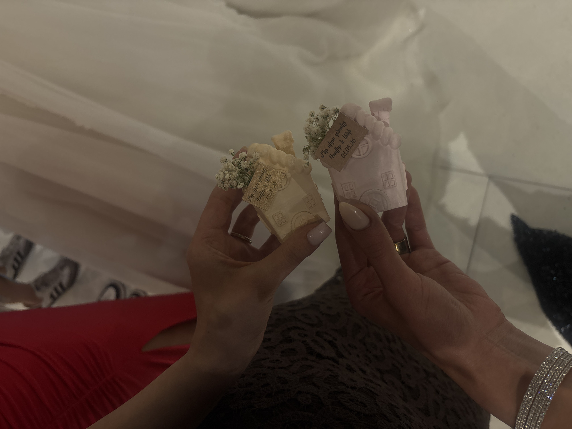 The image shows two hands holding small house-shaped objects, likely wedding favors. One house is cream-colored, and the other is pink. Each house has a small tag with the text "Մեր սիրո տնակը, Դավիթ և Անի, 03.05.26", which translates to "Our Love House, Davit and Ani, 03.05.26", indicating the names of a couple and a date. Both houses are adorned with small white flowers. The person on the left is wearing a red dress, and the person on the right is wearing a diamond bracelet. The location, Masis, Armenia, cannot be confirmed from the image alone.