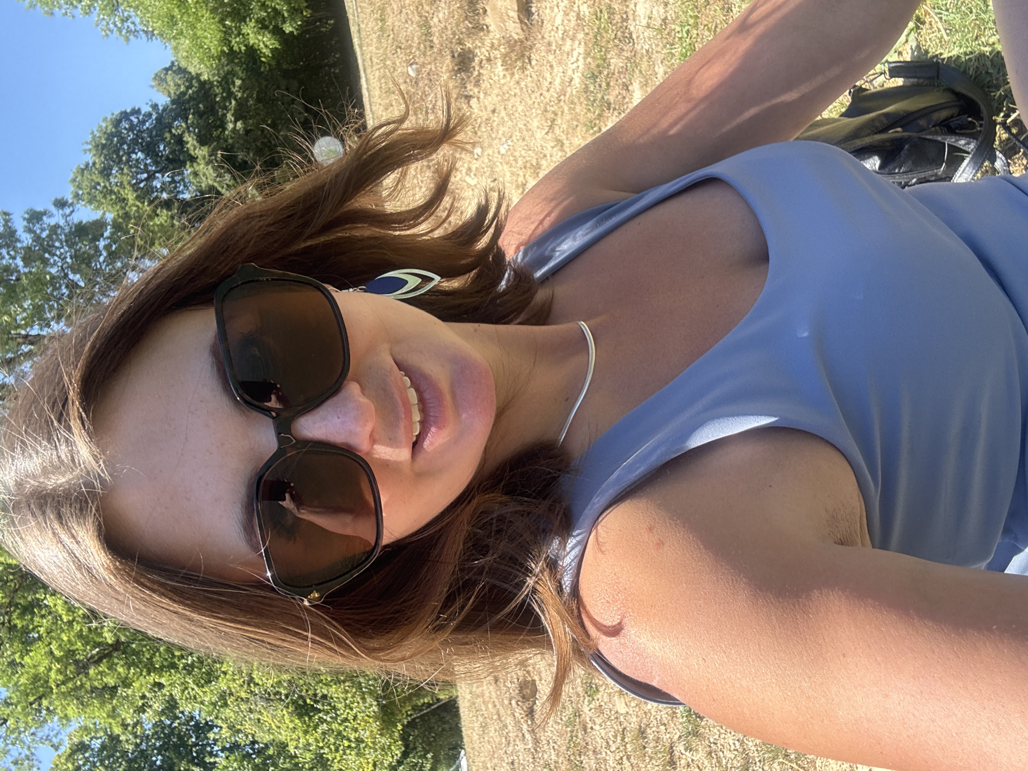 A woman with brown hair and sunglasses is pictured outdoors, seated on dry grass. She is wearing a light blue sleeveless top and a silver necklace. Her right arm is visible, and she appears to be holding a phone, which is reflected in her sunglasses. Behind her, trees and a clear blue sky are visible, suggesting a warm, sunny day. The background appears to be a rural or park setting, possibly in the Coteaux du Lizon region of France.