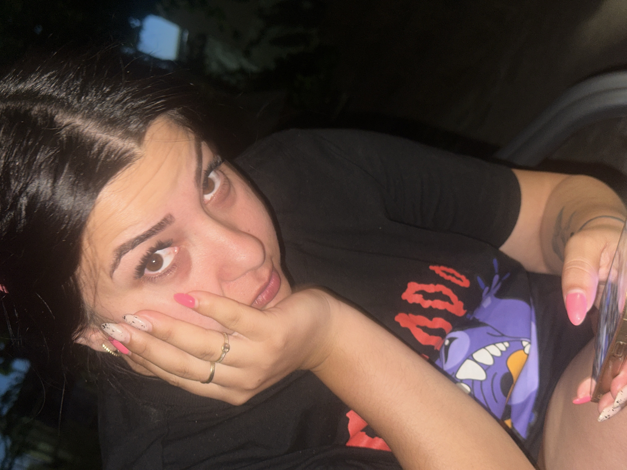 A medium shot captures a young woman in Skala, Bulgaria, resting her face on her hand and looking at a smartphone held in her other hand. The woman has dark hair, is wearing a black t-shirt with a purple cartoon character and red lettering, and has bright pink painted nails. She wears two rings on her right hand. The background is dark and out of focus, with some foliage visible. The lighting appears to be dim, suggesting it is nighttime.