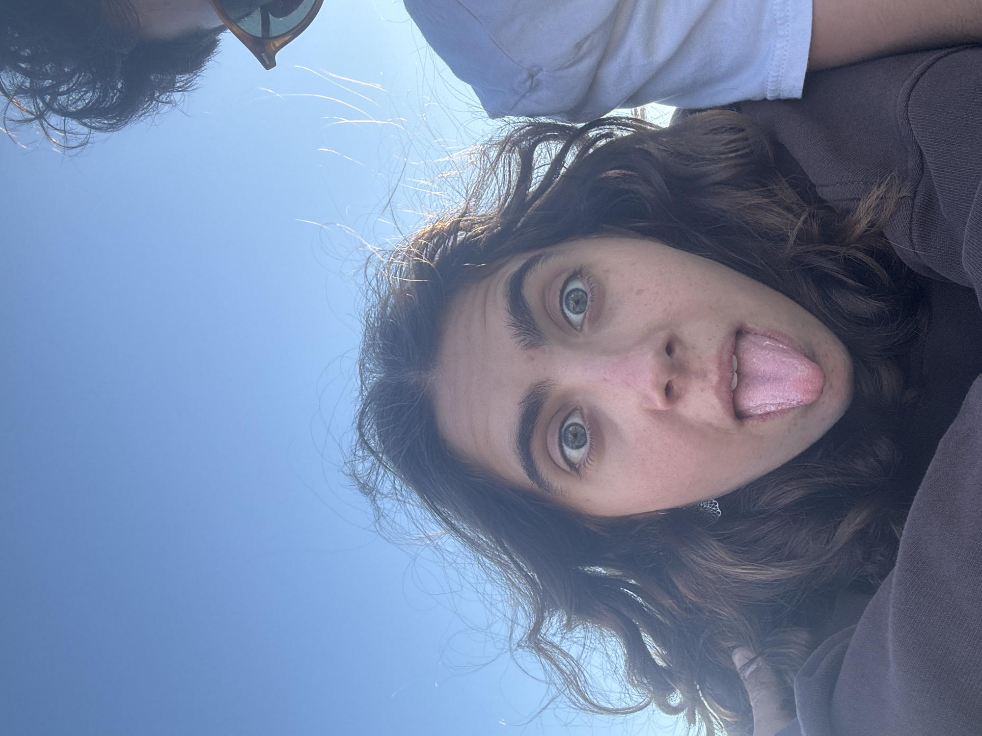 A vibrant, low-angle selfie captures a young woman looking directly at the camera with a playful expression, featuring wide, light-colored eyes and her tongue extended. She has long, dark, wavy hair, brightened by sunlight, and a light complexion with visible freckles. She is wearing a dark brown or grey long-sleeved top, and a small, ornate earring can be seen on her right ear.
Partially visible at the upper left is another person, out of focus, showing dark curly hair and sunglasses, suggesting they are also part of the shot. This person appears to be wearing a white t-shirt. The entire background is a clear, bright blue sky, indicative of a sunny outdoor setting with no clouds. The strong sunlight suggests it is daytime, likely midday or early afternoon. The image conveys a spontaneous, energetic, and lighthearted mood. While no specific visual cues for Tel-Aviv, Israel are present in the photo itself, this is the provided location context. There is no visible text in the image.