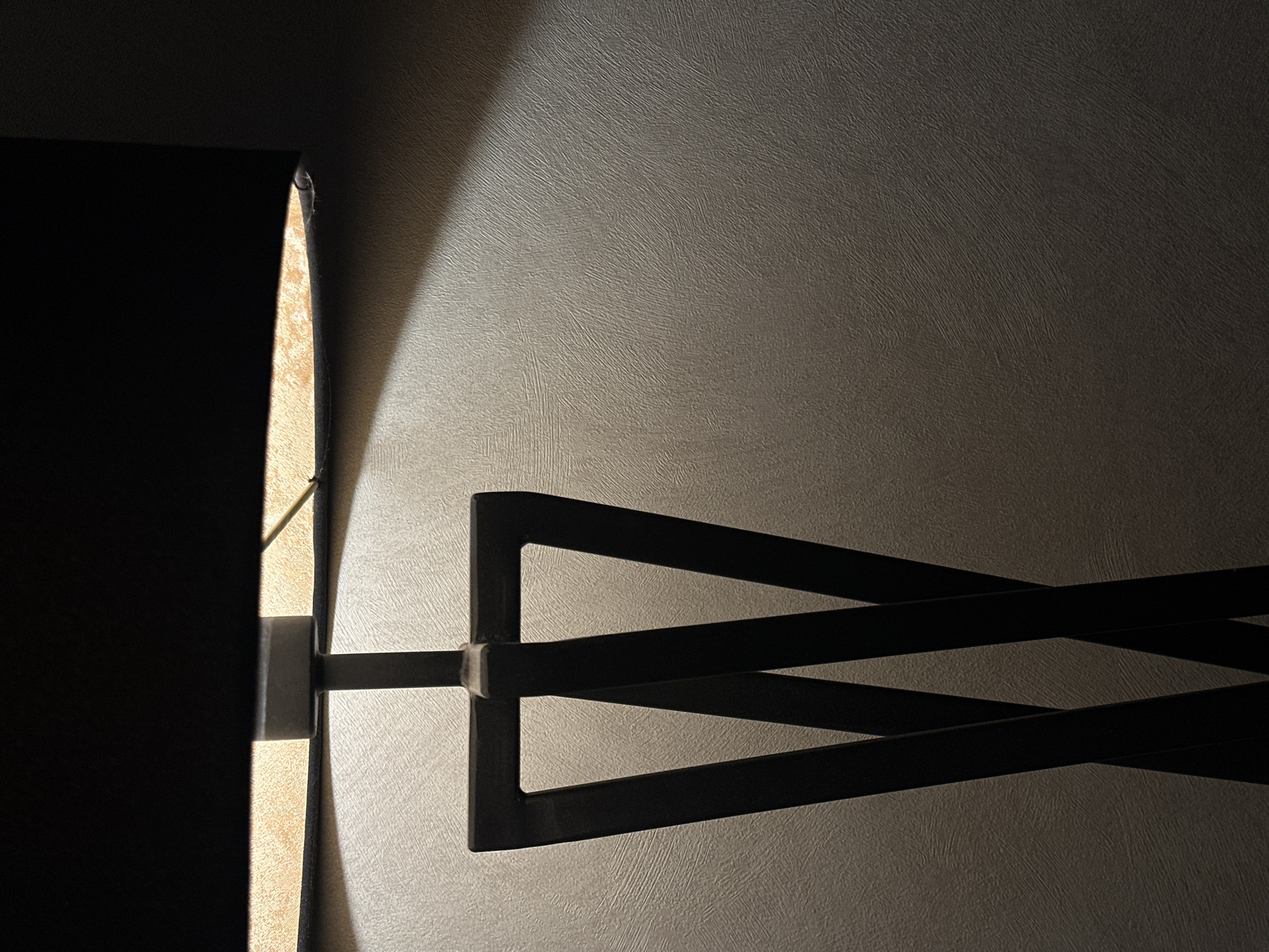 A dimly lit, close-up indoor photograph shows a wall-mounted lamp with a black shade. The lamp extends from the wall via a black, geometric arm consisting of two connected rectangles. The wall has a textured, neutral-toned surface. The lamp shade is partially illuminated, emitting a warm glow that casts a soft, angular shadow on the wall. The setting is unknown, there is no evidence to suggest that this image was taken in the city of Chaikovsky, Russia.