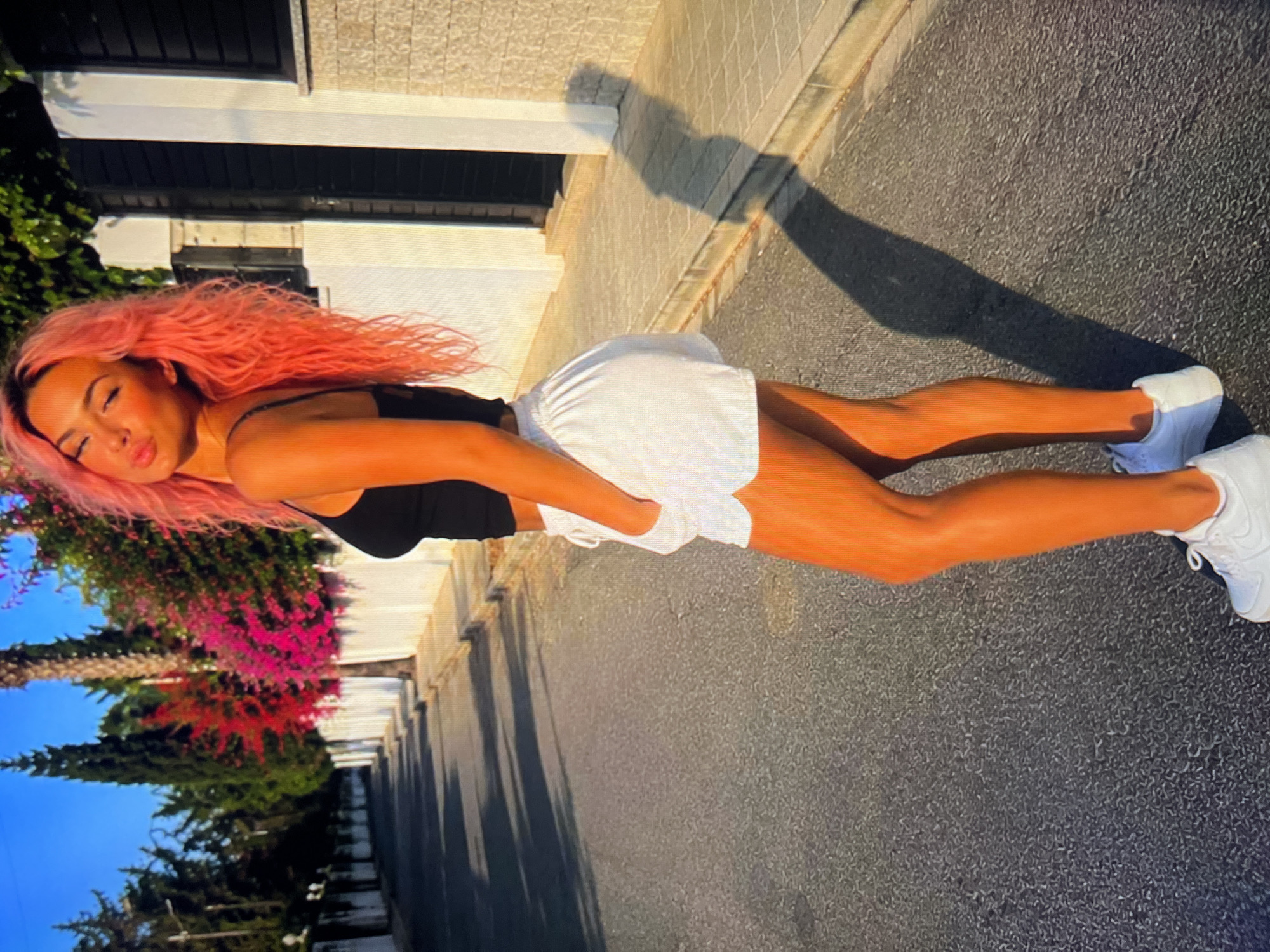 A young woman with bright pink hair is standing on an asphalt street in front of a white fence with flowering bushes. She is wearing a black tank top and white shorts, and white sneakers. The sun is shining brightly, casting a long shadow of the woman on the street. The street appears to be in a residential area, with houses and trees visible in the background.