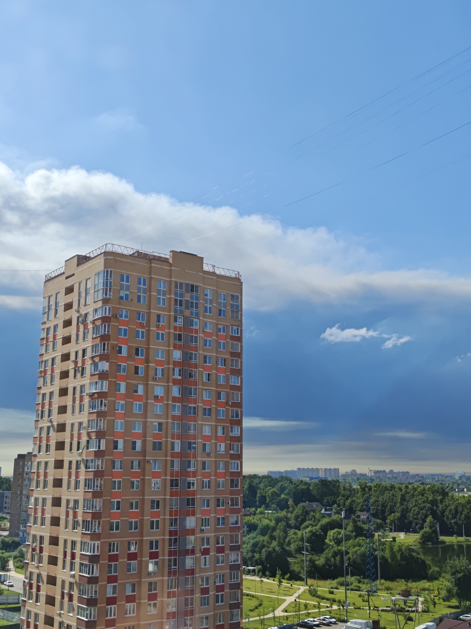 The image depicts an urban landscape in Shcherbinka, Russia, dominated by a tall, modern apartment building with a light brown facade and orange accents around the windows. The building features numerous windows and balconies, suggesting a residential purpose. In the background, a lush green forest with a body of water is visible, interspersed with smaller buildings and a distant cityscape. The sky is a mix of bright blue with scattered white clouds and darker, more dramatic storm clouds, hinting at changing weather conditions. Several power lines are visible stretching across the sky, and there are also faint contrails from aircraft. The foreground shows a neatly manicured park or recreational area with paved pathways, grassy areas, parked cars, and playground equipment. Another apartment building is partially visible to the left of the main subject. There are no people visible in the image.