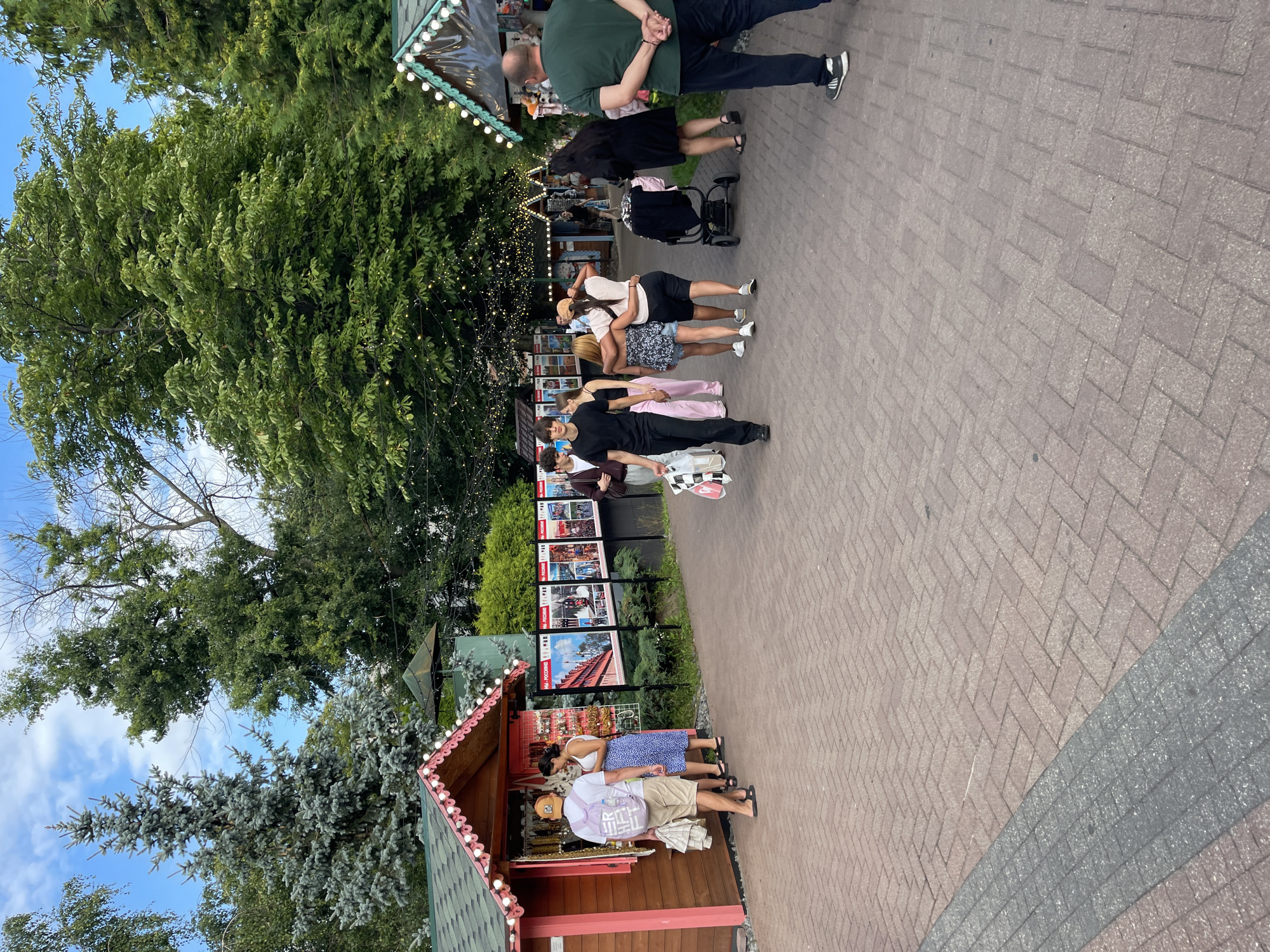 In an outdoor, paved pathway area with brick-like paving, a diverse group of people are walking, browsing, and conversing. On the left, two individuals are near a small, pink-accented wooden booth, one of whom is wearing a backpack and looking at items displayed in the booth. To the right of this booth are several posters displaying images, suggesting a tourist attraction or event.
Further down the path, a group of young people are walking together, some with arms around each other. A stroller is visible near this group. On the far right, a man in a green shirt and dark pants stands with his hands behind his back, facing away from the camera.
The environment is lush with trees overhead, some of which are decorated with string lights. The sky is visible and appears partly cloudy. Two more similar booths, one with a green roof and pink trim, are visible in the background, also adorned with lights. The overall scene suggests a lively outdoor public space, possibly a park, fairground, or a street lined with vendors.
This location appears to be in the city of Kaliningrad, Russia.