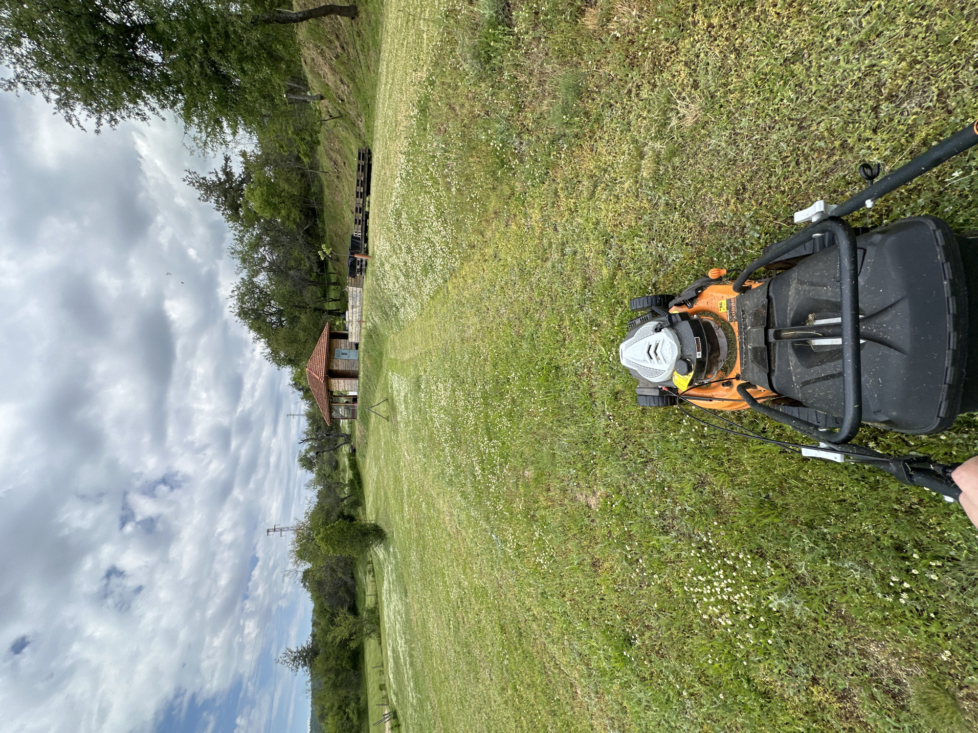 The image shows a person mowing a grassy field in Mrachenik, Bulgaria. The lawnmower is orange and black. A path of cut grass is visible in front of the lawnmower, leading towards a small building with a red tile roof in the distance. The sky is partly cloudy. Trees border the edge of the field.