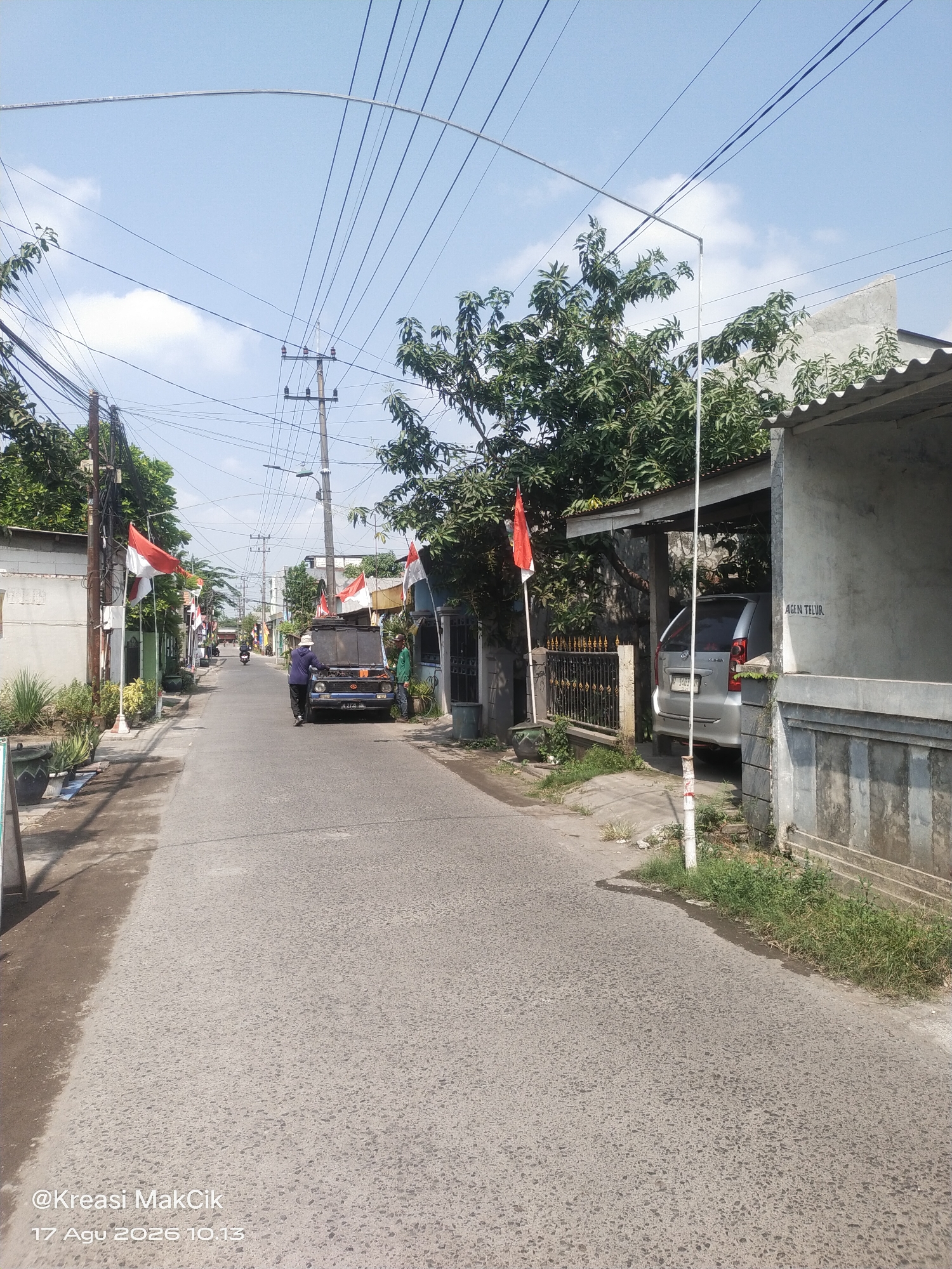 The image depicts a street scene in Sambi Bulu, Indonesia, likely on or around Indonesia's Independence Day, August 17th, as indicated by the watermark "@Kreasi MakCik 17 Agu 2026 10.13". The street is paved and runs through a residential area with houses on both sides. Several red and white Indonesian flags are prominently displayed along the street, attached to poles and railings.
On the left side of the street, there are houses with some vegetation, potted plants, and what appear to be decorative pots. Utility poles with numerous electrical wires crisscross overhead, creating a dense network against the blue sky with scattered clouds.
In the middle of the street, a dark-colored pickup truck with its hood open is parked. Two individuals are near the truck. One person, wearing a blue shirt and a white hat, is standing next to the open hood. Another person, in a green shirt, is standing to the right of the truck. Further down the street, a motorcycle is visible, suggesting ongoing traffic.
On the right side of the street, a silver minivan is parked under a carport attached to a house. A sign on the wall of the house reads "AGEN TELUR", which translates to "Egg Agent". The house also has a concrete fence. The environment appears to be a typical Indonesian neighborhood with a mix of residential buildings and public infrastructure. The overall impression is of a calm day in a community decorated for a national celebration.
