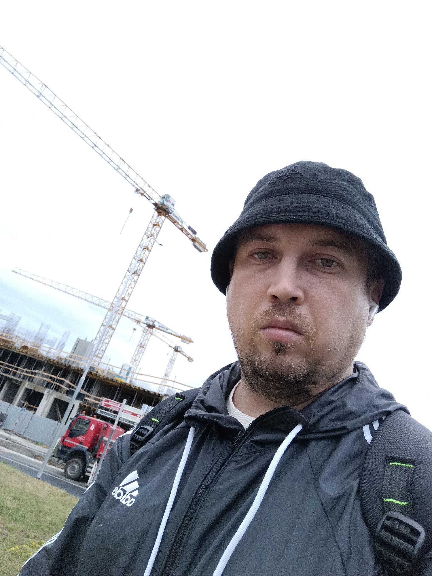 The media file is a selfie of a man in his 30s wearing a black bucket hat and a black and white Adidas jacket, with a backpack on. He has short brown hair and stubble, and he is wearing a white wireless earbud in his right ear. His expression is neutral.
The background shows a construction site with several large cranes and unfinished buildings. A red truck is visible on the left side of the image. The sky is overcast, suggesting a cloudy day. The location is identified as Prague, Czechia.
The man appears to be outdoors at a construction site, possibly on his way to or from work, or taking a break. The overall impression is of a man in an urban environment engaged in or observing a construction project. The image seems to be taken during daylight hours, given the visibility of the scene.