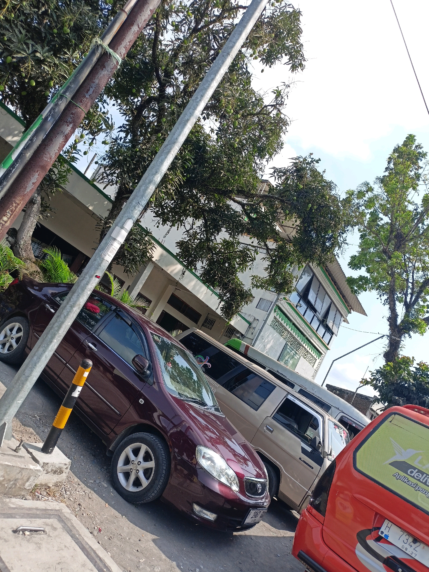 In Payaman, Indonesia, on a sunny day, a street scene unfolds. A maroon sedan is parked on the left side of the frame, its front tire angled towards the camera. To its right, a beige van is parked, followed by a red vehicle, only partially visible, featuring a yellow sticker with green text that appears to be a delivery company logo. Behind the vehicles, a white building with green accents and patterned window designs stands, with trees, some bearing fruit, in front and to the side of it. Utility poles, one rusty and one silver, are positioned in the foreground, intersecting the view of the street and buildings. The ground is a mixture of asphalt and dirt, with concrete elements in the lower left corner. The sky is bright and partially obscured by tree branches.