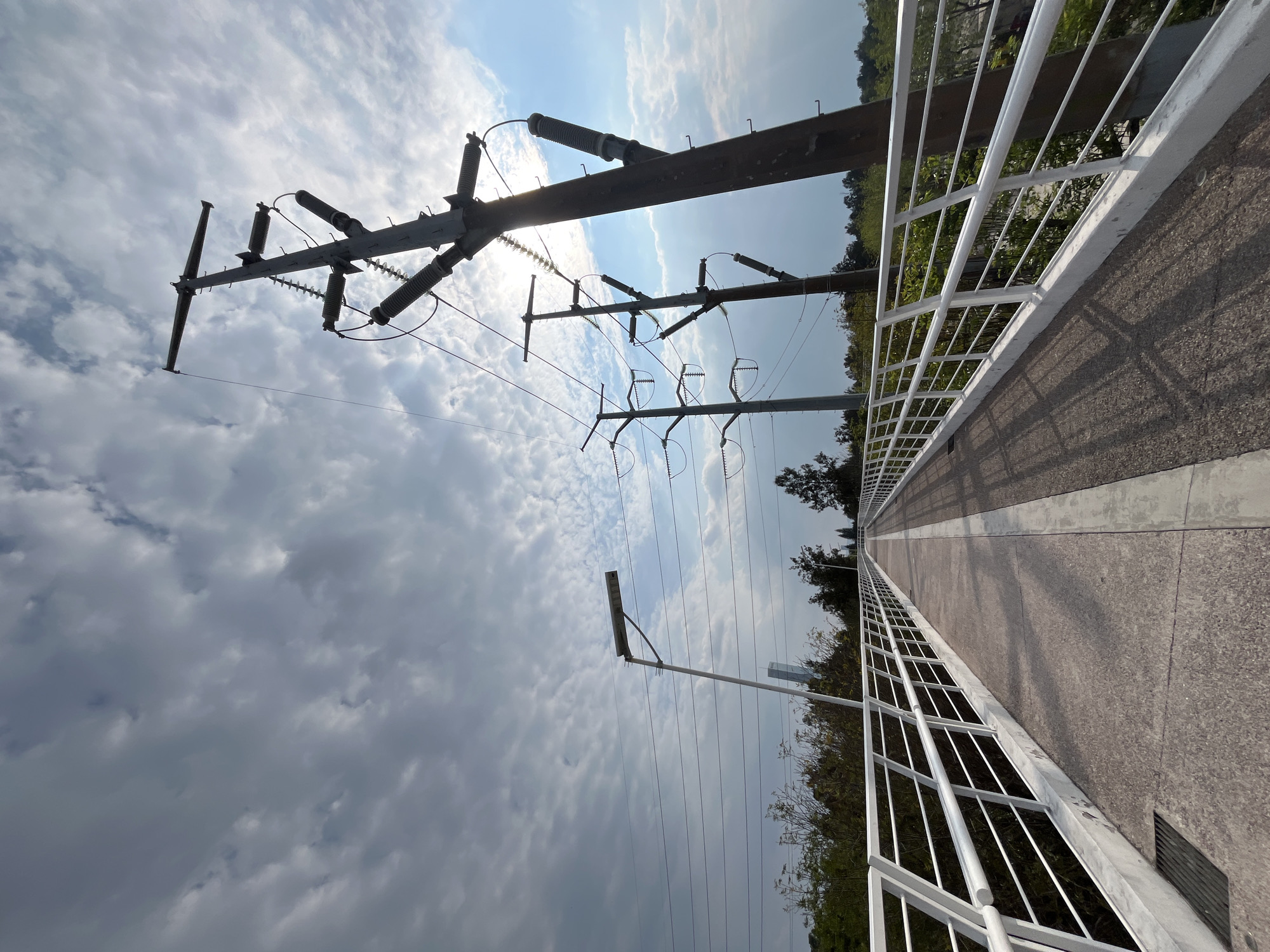 The image shows a pedestrian bridge with white metal railings, leading into the distance under a cloudy sky. Electrical power lines and wooden support structures run alongside the bridge. The surface of the bridge appears to be concrete or asphalt. Vegetation is visible on either side of the bridge. A street light is also visible. There is no evidence to confirm this image is from Puebla, Mexico.