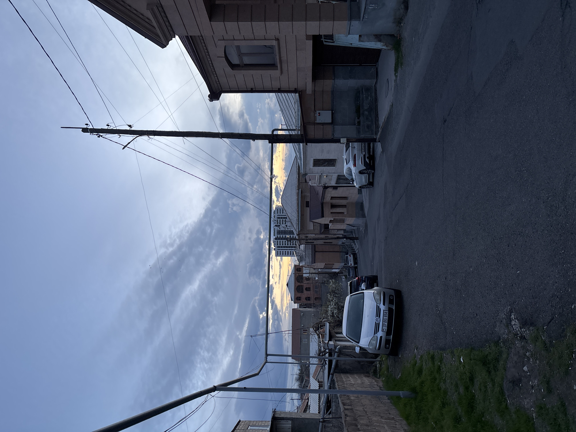 This image depicts a street in Yerevan, Armenia, with a view looking downhill between buildings. The sky is overcast with hints of a sunset, and power lines stretch across the scene. Parked cars are visible on the right side of the street, a white Volkswagen Golf being the most prominent. The road is asphalt, and various residential buildings line the street. In the distance, taller buildings can be seen.