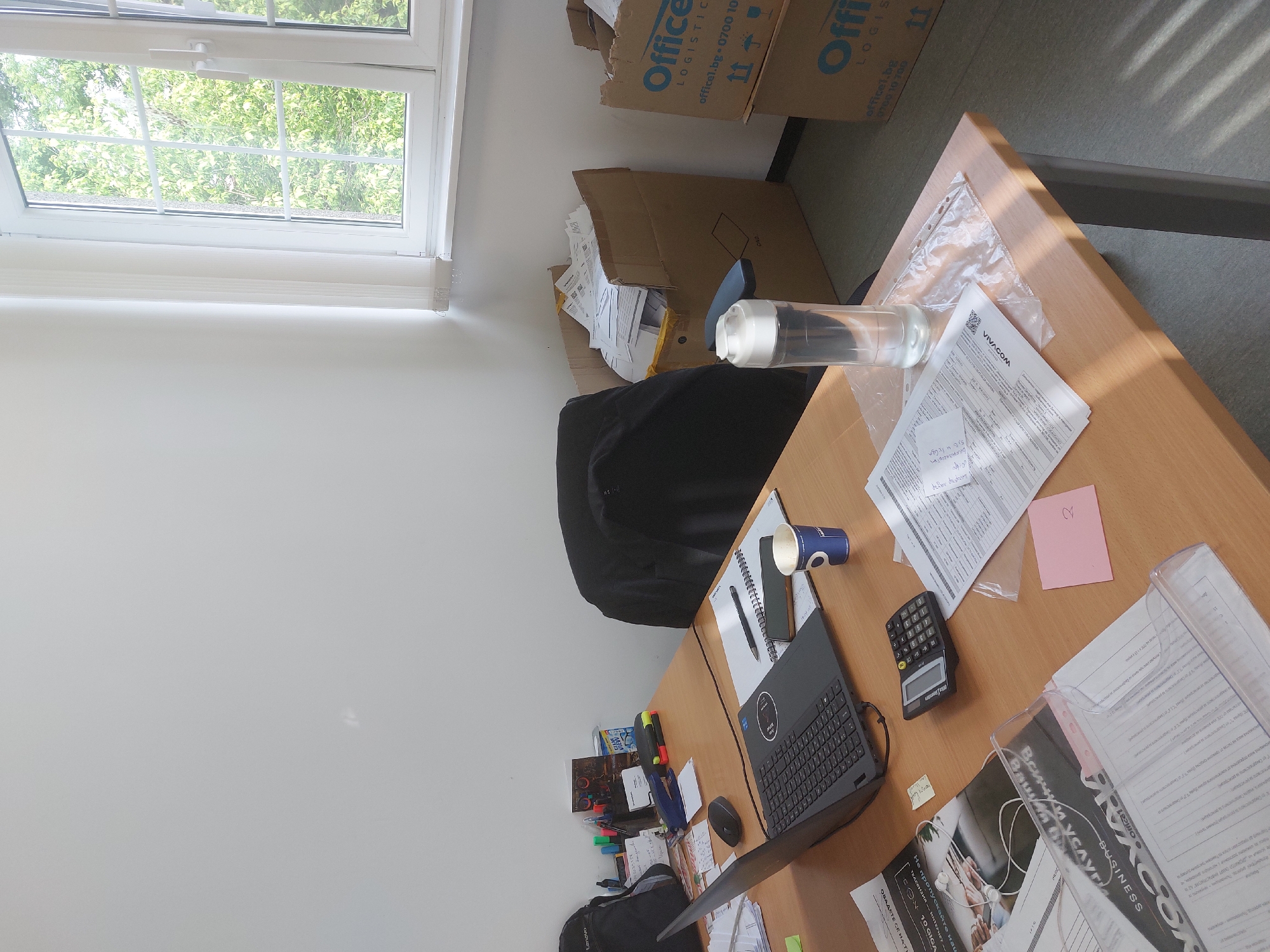 The image depicts a cluttered desk in what appears to be an office environment, possibly in Burgas, Bulgaria. The desk is light brown and has various items on it, including a black laptop with a Lenovo logo, a black calculator, a blue paper cup, a spiral notebook, a black pen, and a black smartphone. Documents with what seems to be "VIVACOM" printed on them and a pink sticky note with a number "2" are on the desk. There are also office supplies, such as pens and highlighters. There is a box from "Office Logistics" which is located in Burgas. A black office chair is positioned near the desk. Natural light comes through a window in the upper left corner of the image, and boxes are stacked near the window. A small amount of the background floor is visible and is grey.