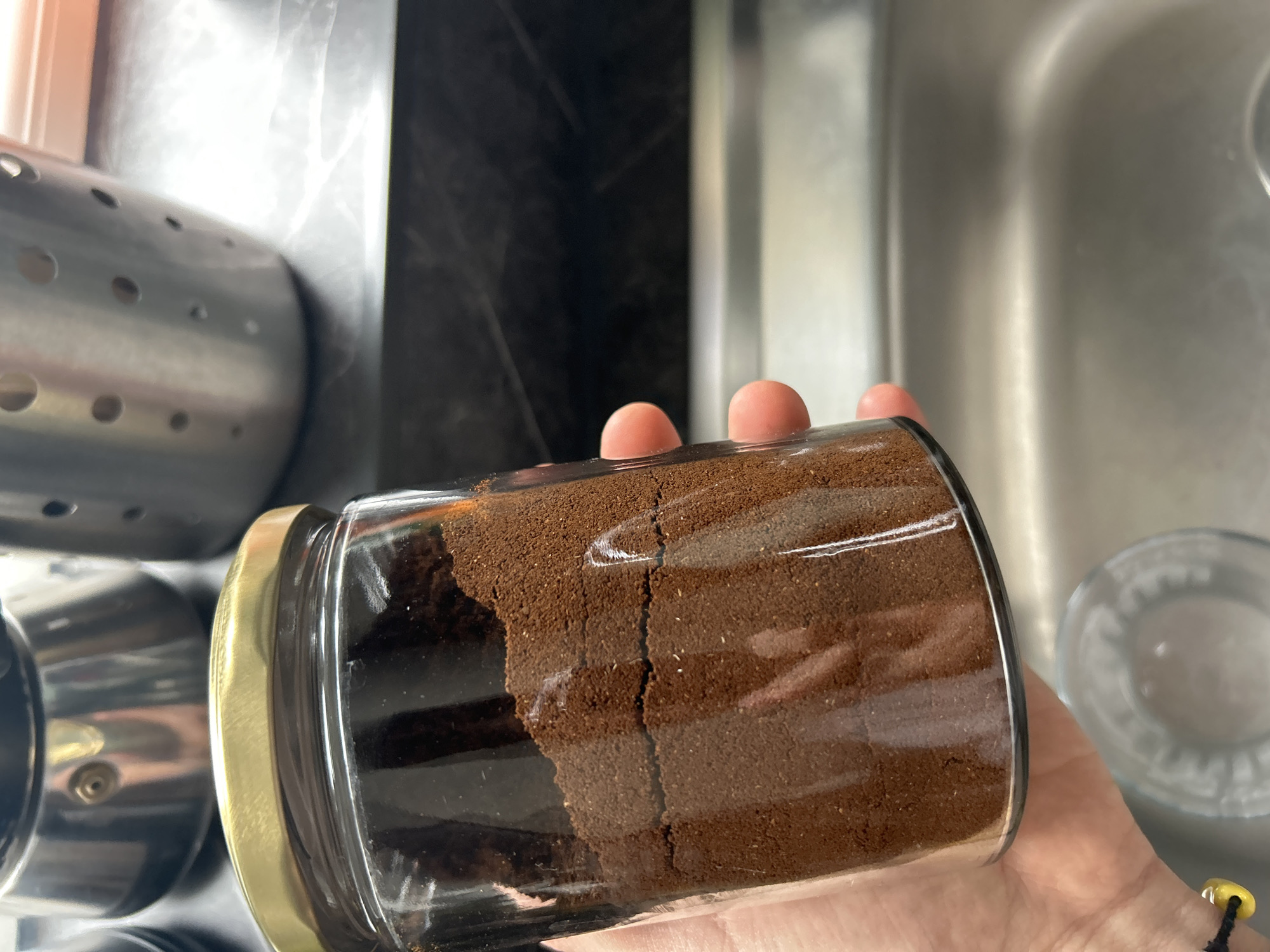 A person is holding a glass jar filled with coffee grounds. The jar has a gold lid. The background includes a metal utensil holder and a kitchen sink. The location cannot be determined from the image.